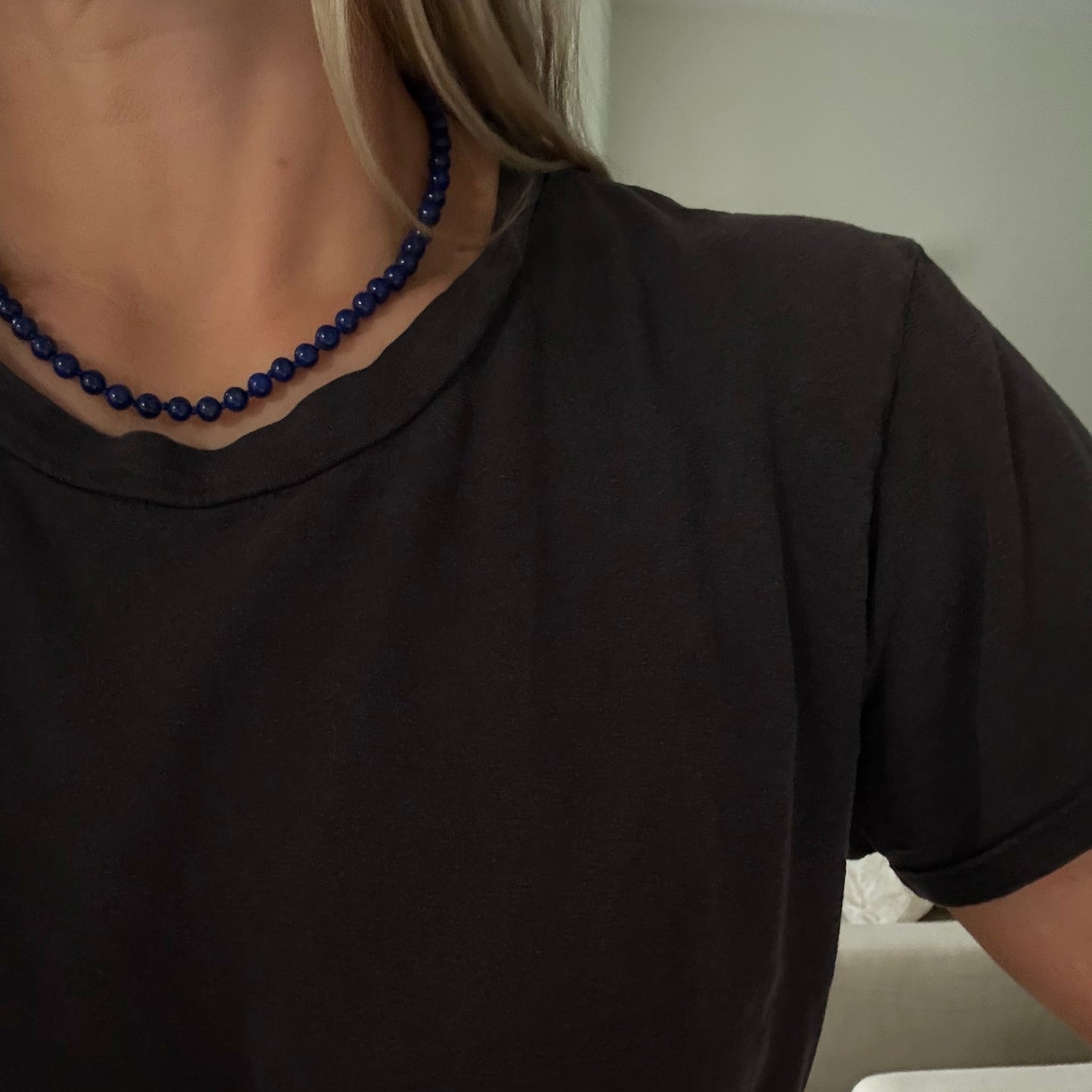 A close-up of a person wearing a dark charcoal gray crewneck t-shirt and a dark blue beaded necklace, with a white armchair visible in the background.