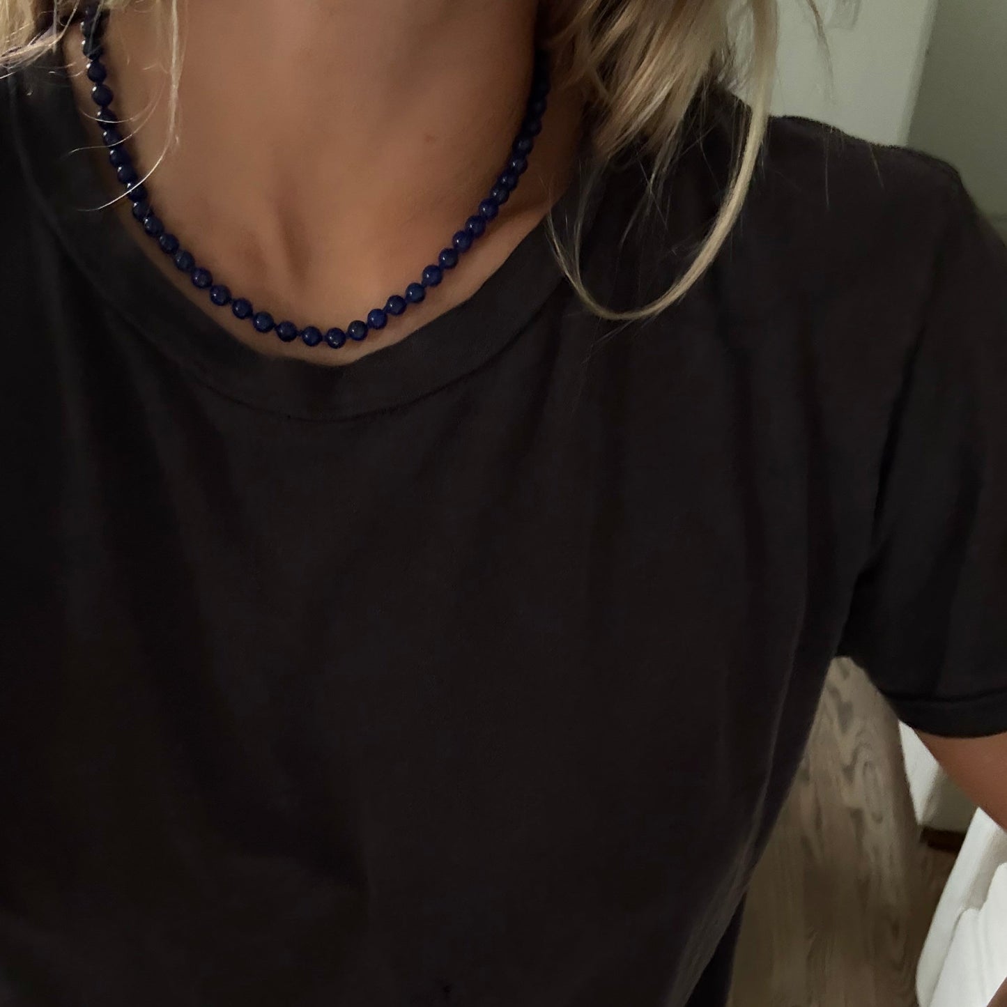 A close-up of a person wearing a dark charcoal gray t-shirt and a dark blue beaded necklace, with blonde hair visible.