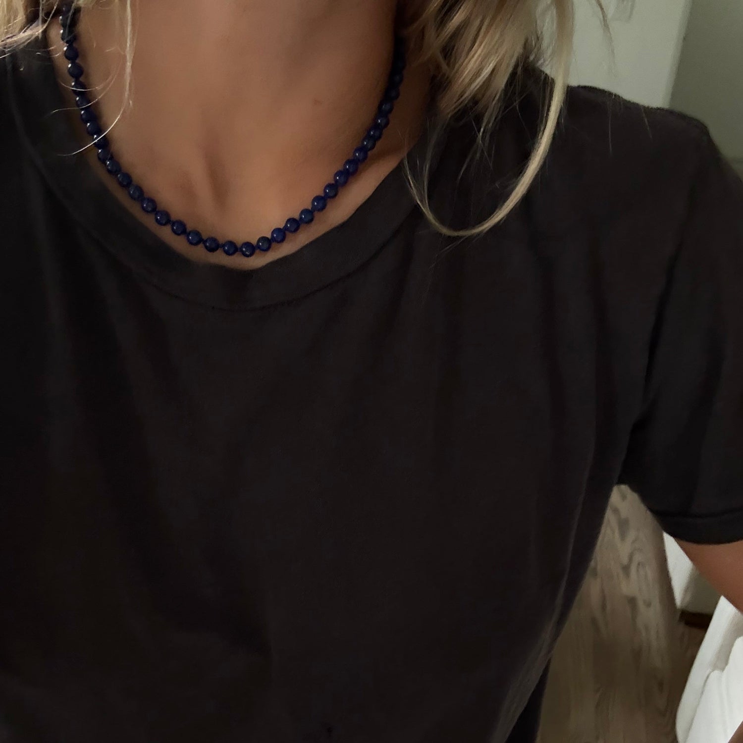 A close-up of a person wearing a dark charcoal gray t-shirt and a dark blue beaded necklace, with blonde hair visible.