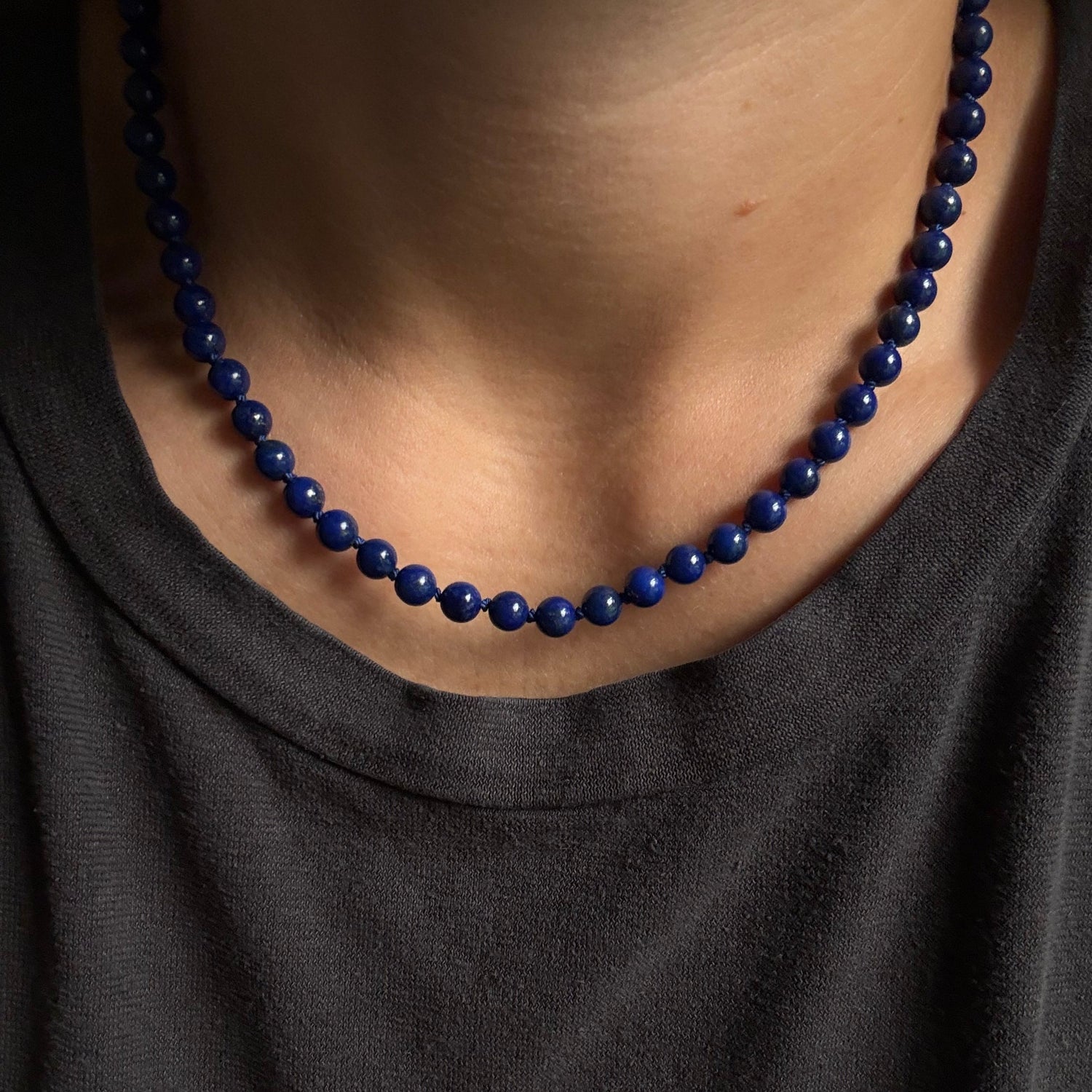 A close-up of a person wearing a dark charcoal gray t-shirt and a dark blue beaded necklace.