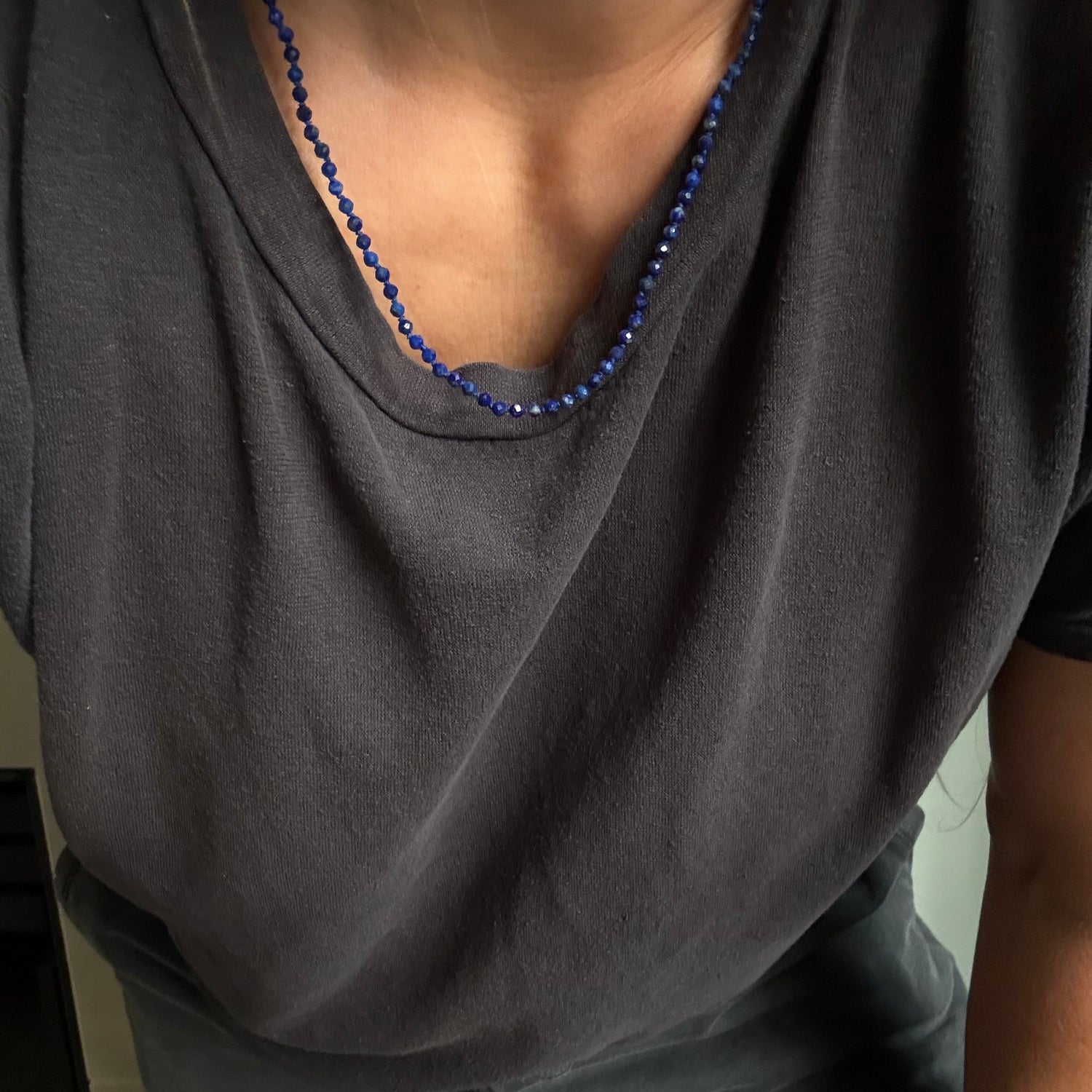 A close-up of a person wearing a dark charcoal gray t-shirt and gray pants, accessorized with a blue beaded necklace.
