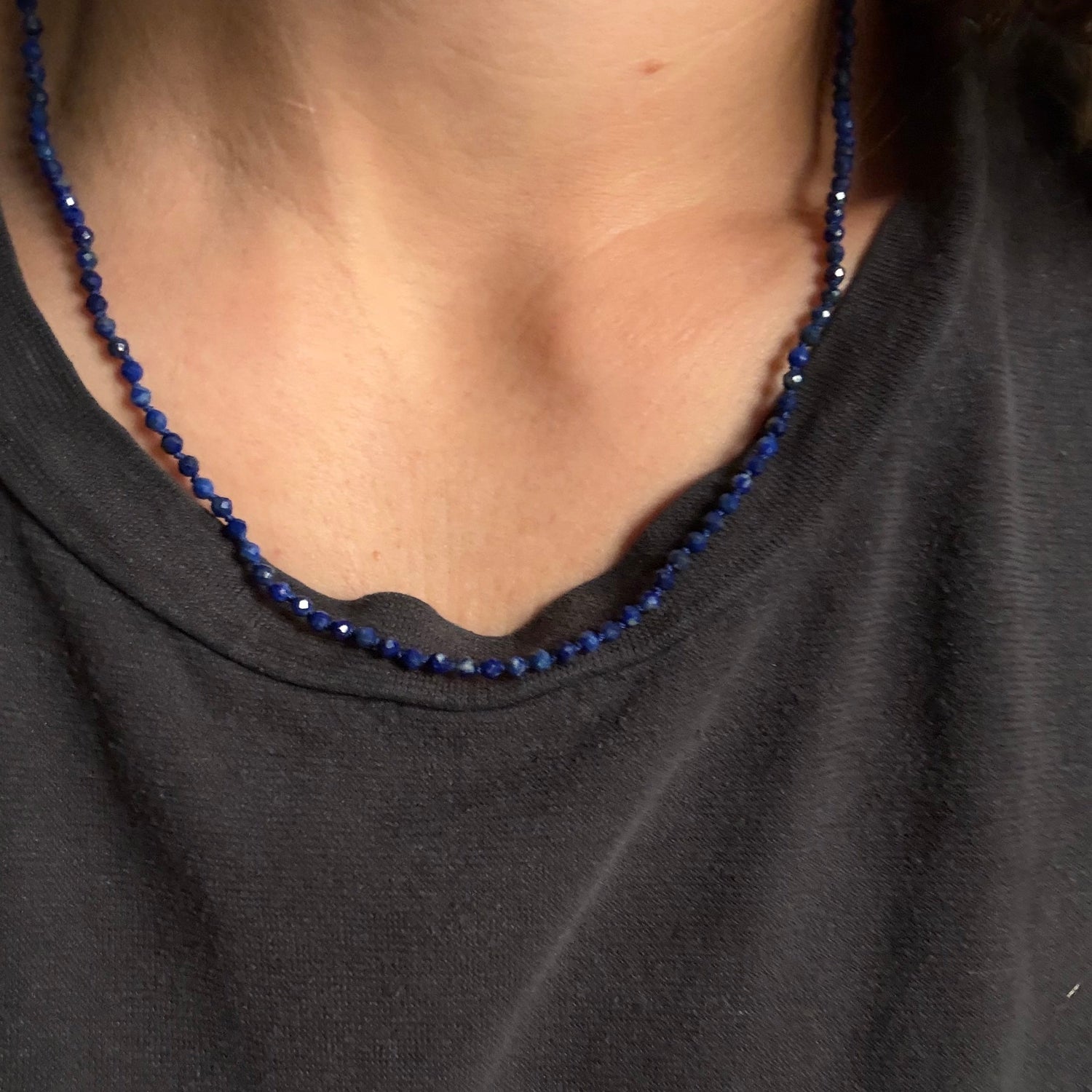 A close-up of a person wearing a dark charcoal gray t-shirt and a beaded necklace made of small, faceted dark blue beads.
