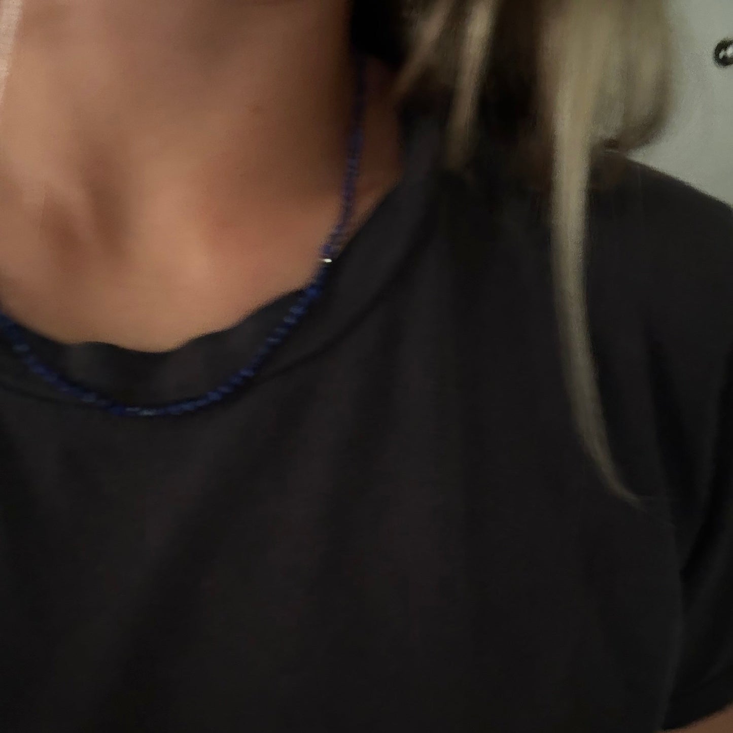 A close-up of a person wearing a dark charcoal gray t-shirt and a thin blue beaded necklace.