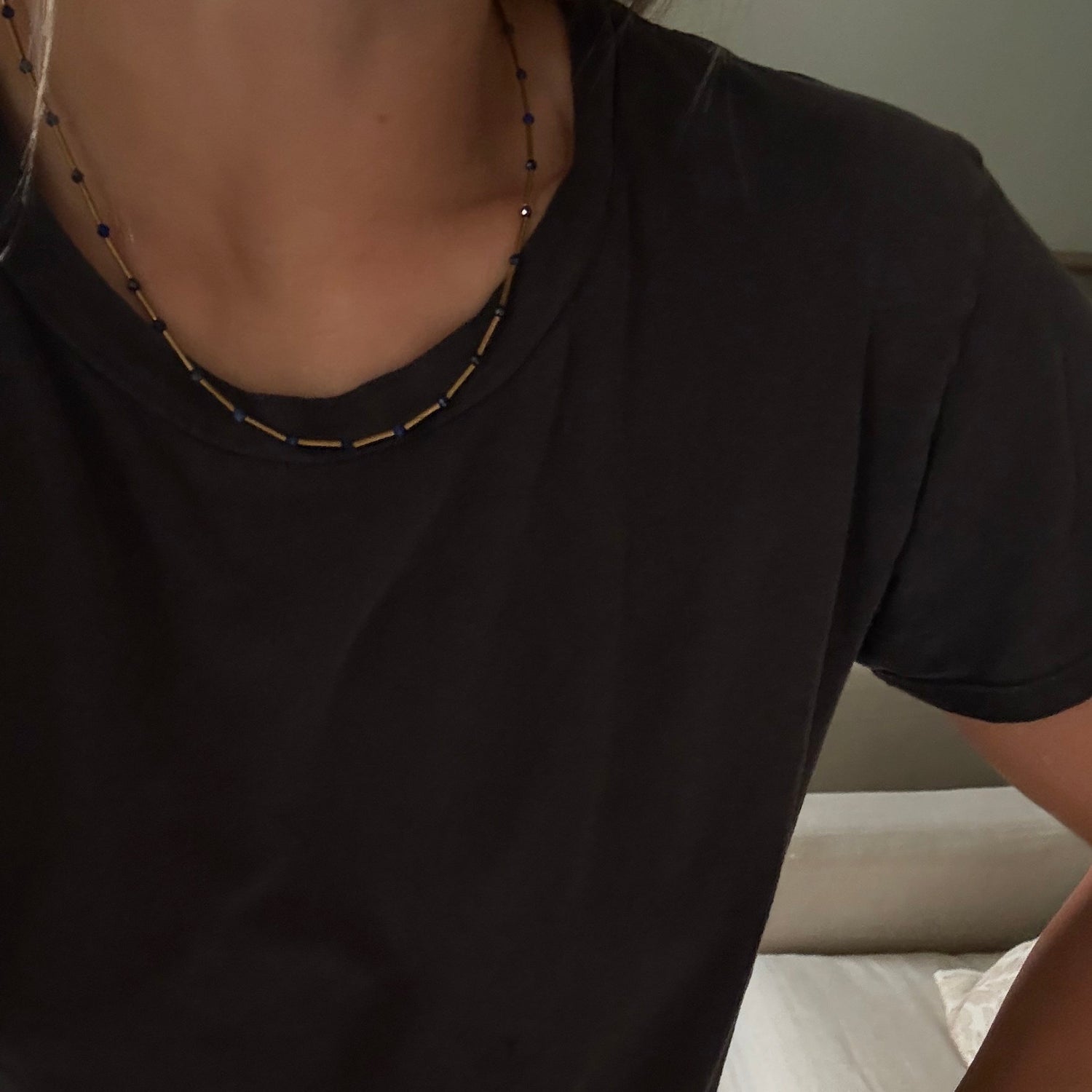 A close-up of a person wearing a black short-sleeve crewneck t-shirt, a beaded necklace, and a smartwatch, standing in front of a bed with white sheets.