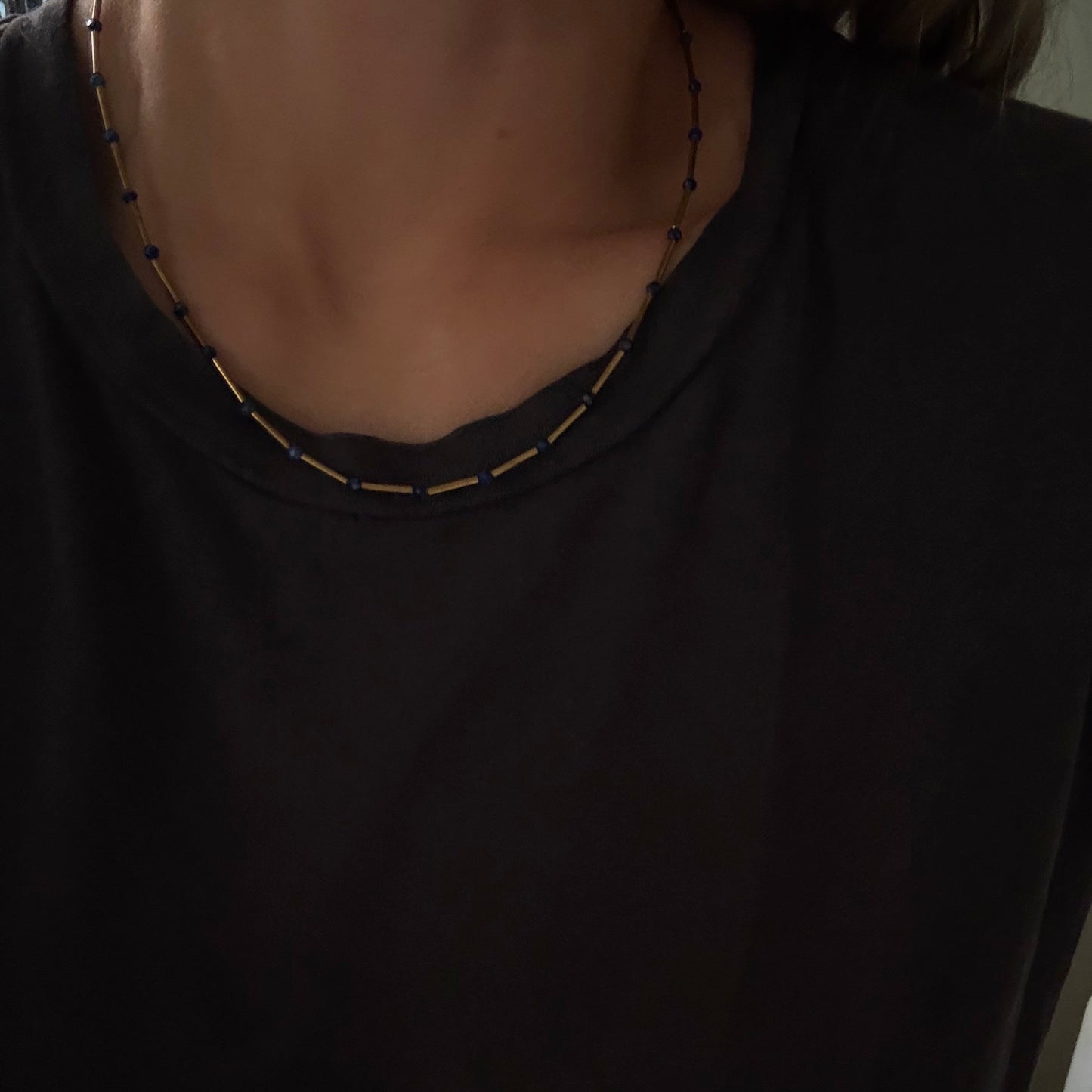 A close-up of a person wearing a black crew-neck t-shirt and a beaded necklace with small gold and dark beads.