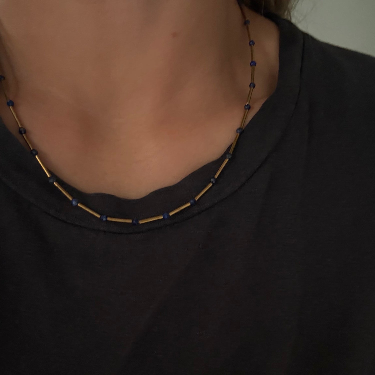 A close-up of a person's neck wearing a beaded necklace with alternating dark blue beads and small gold-toned tube beads, paired with a black t-shirt.