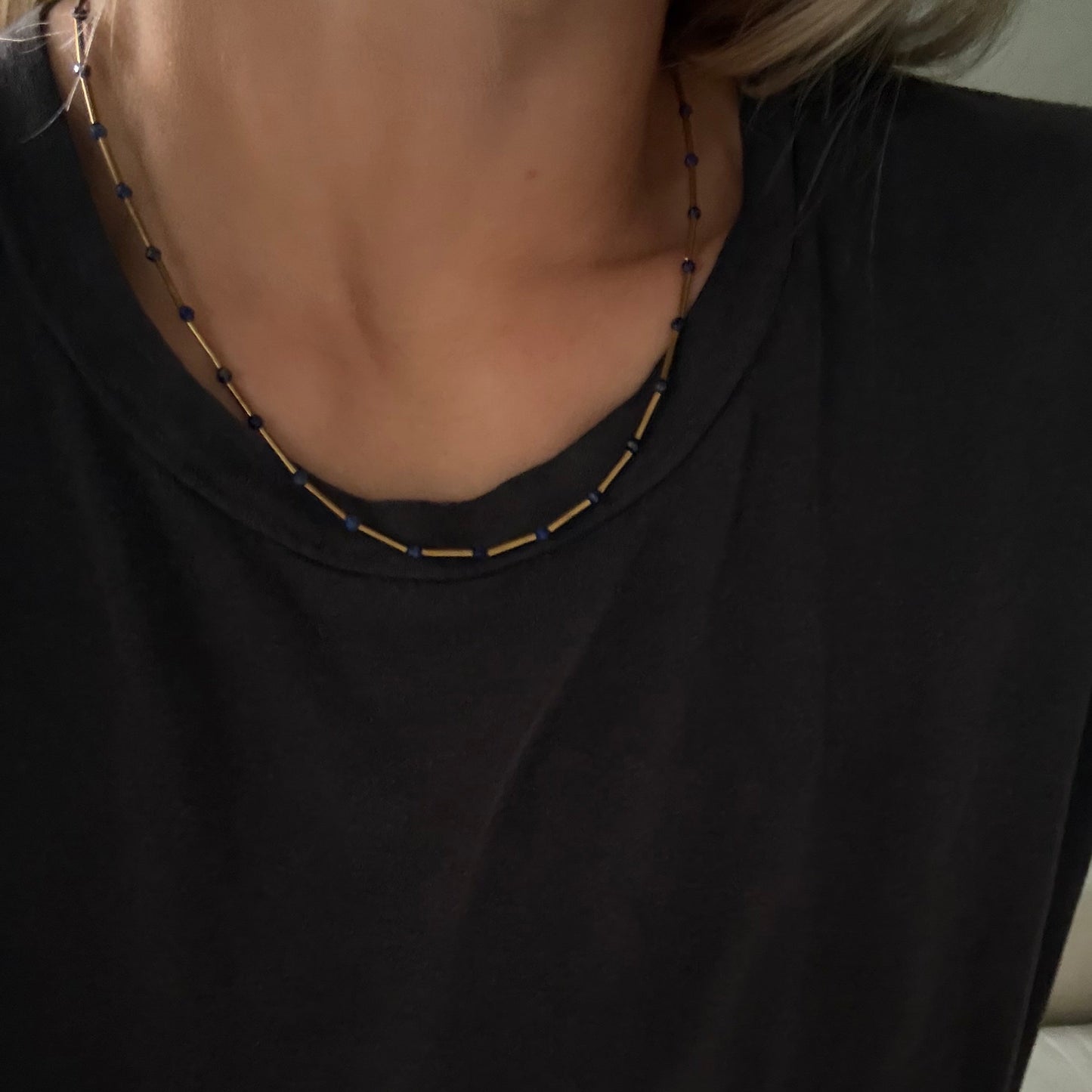 A close-up of a person wearing a black t-shirt and a delicate beaded necklace with gold and dark blue beads.