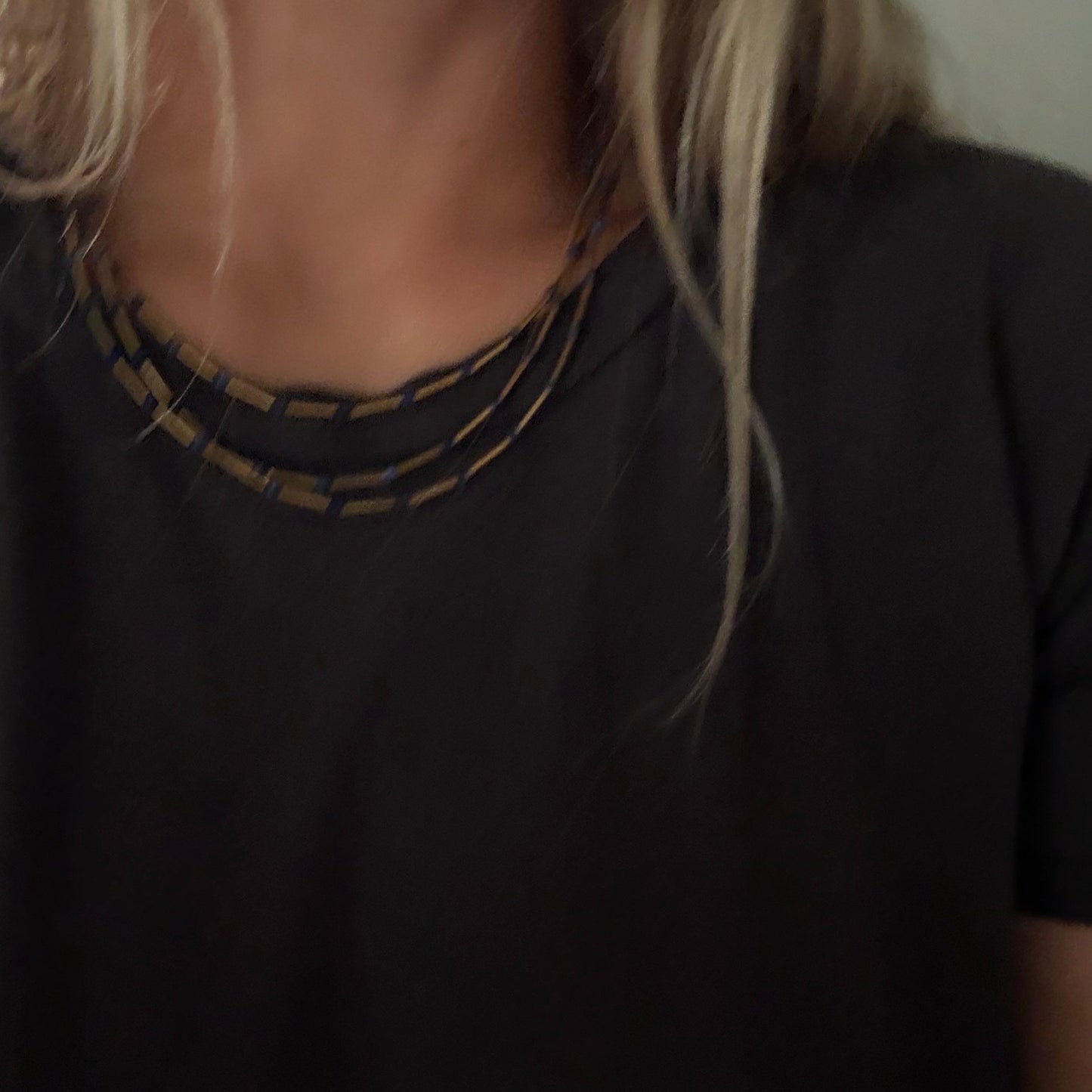 Close-up of a person wearing a black crew-neck t-shirt with a layered geometric beaded necklace in black and gold.