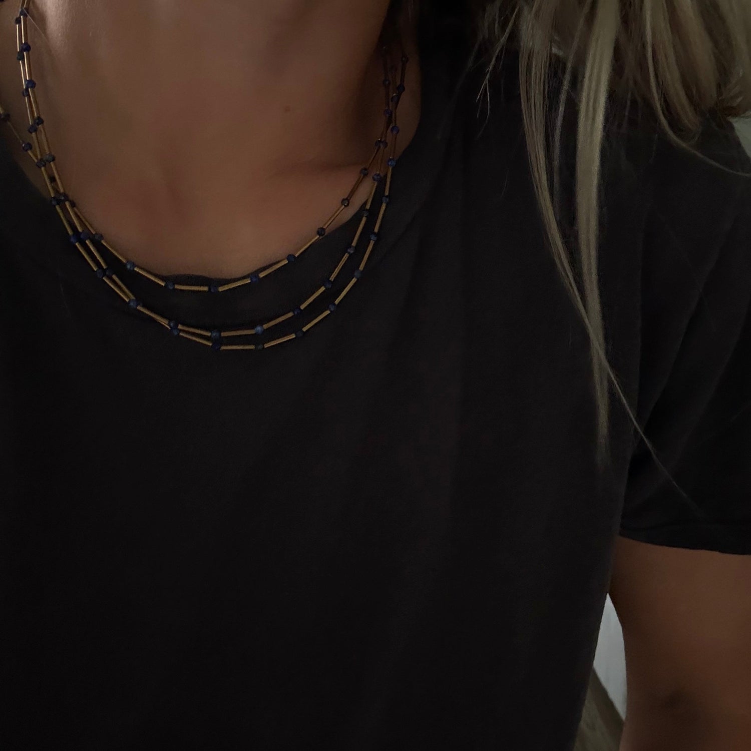 A close-up of a person wearing a layered necklace with small gold and dark blue beads over a black t-shirt.
