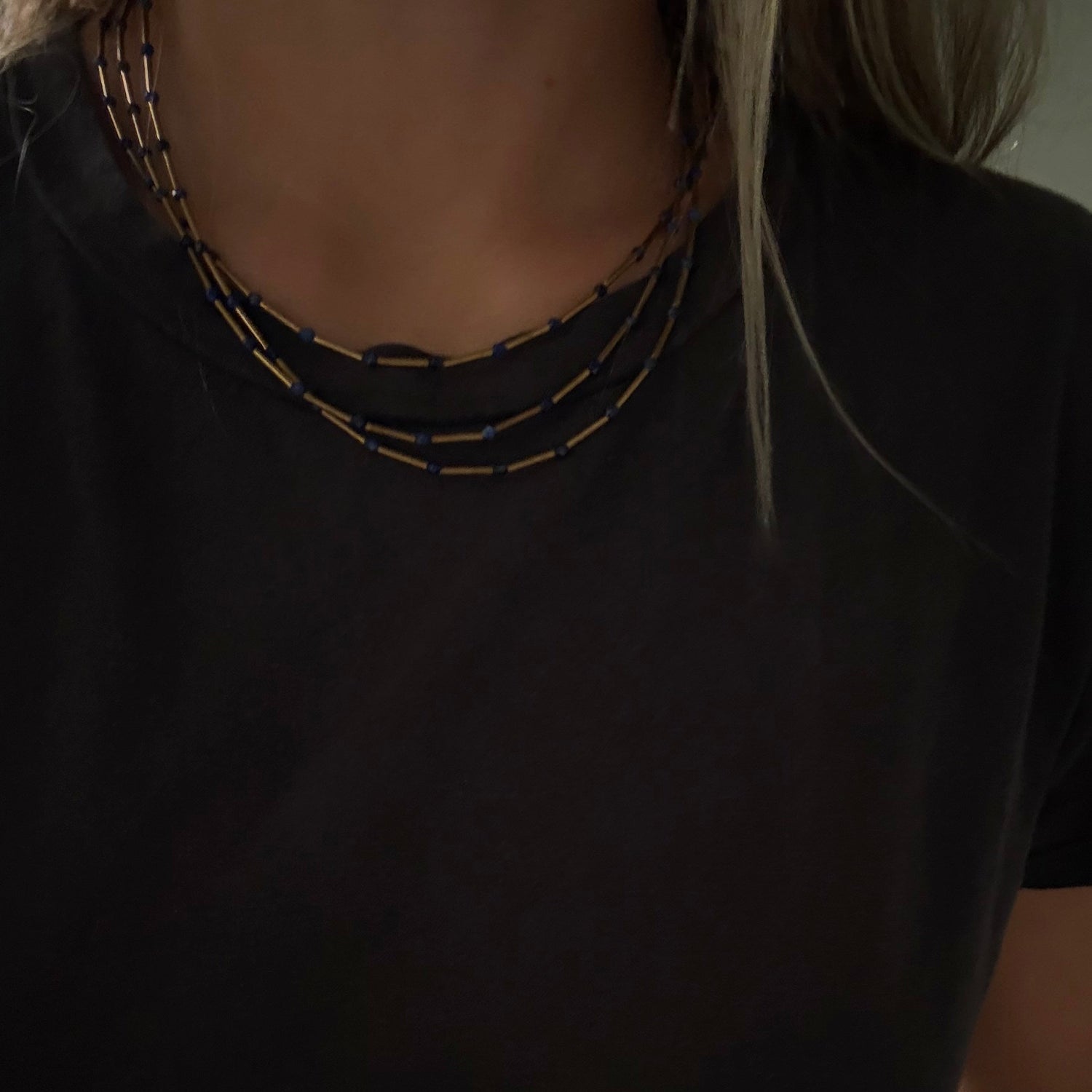 A close-up of a person wearing a black short-sleeve t-shirt and a multi-strand necklace made of small gold-toned and dark metallic beads.