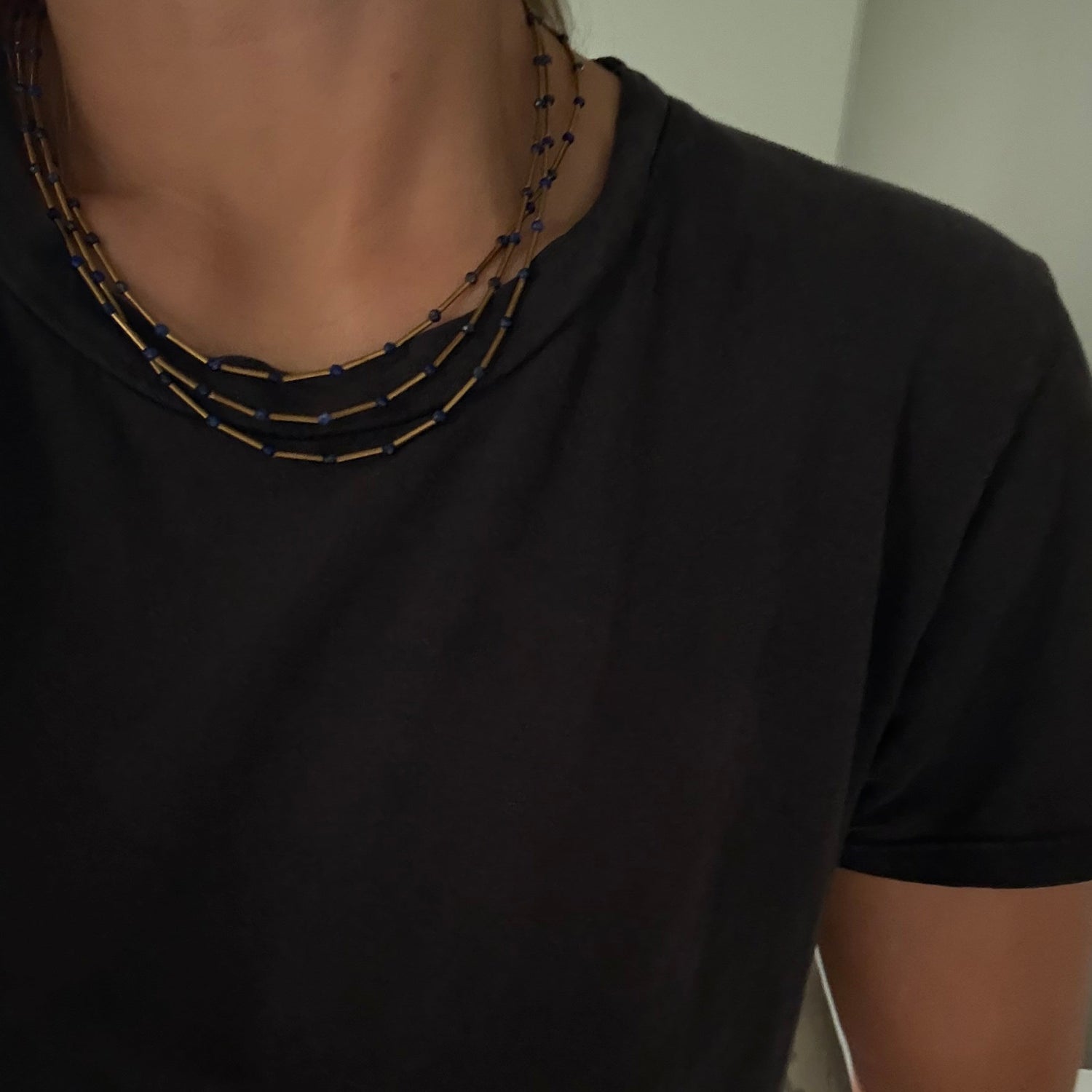 A close-up of a person wearing a layered necklace with small gold and dark blue beads over a black t-shirt.