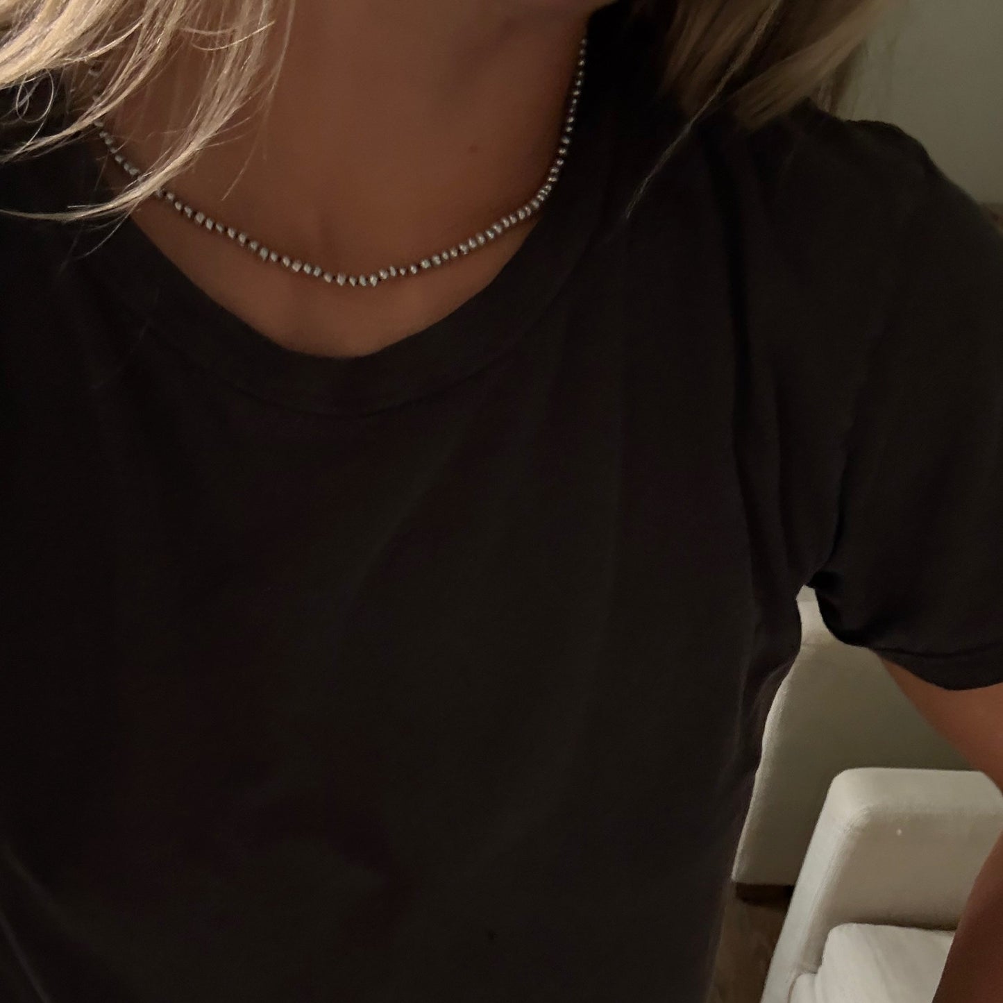 A close-up of a person wearing a delicate gray freshwater pearl necklace with a black t-shirt in a dimly lit indoor setting.