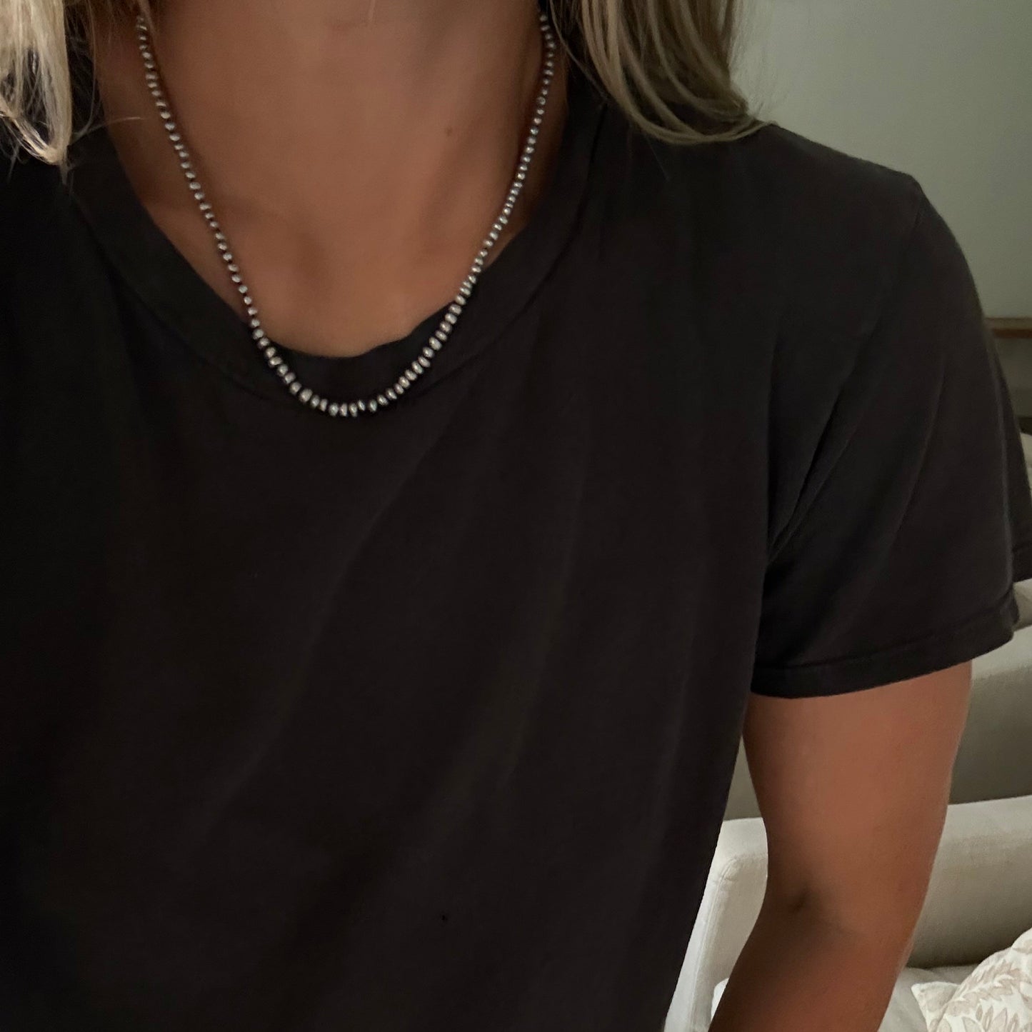 A close-up of a person wearing a gray freshwater pearl necklace  over a black t-shirt, with blonde hair visible at the top.