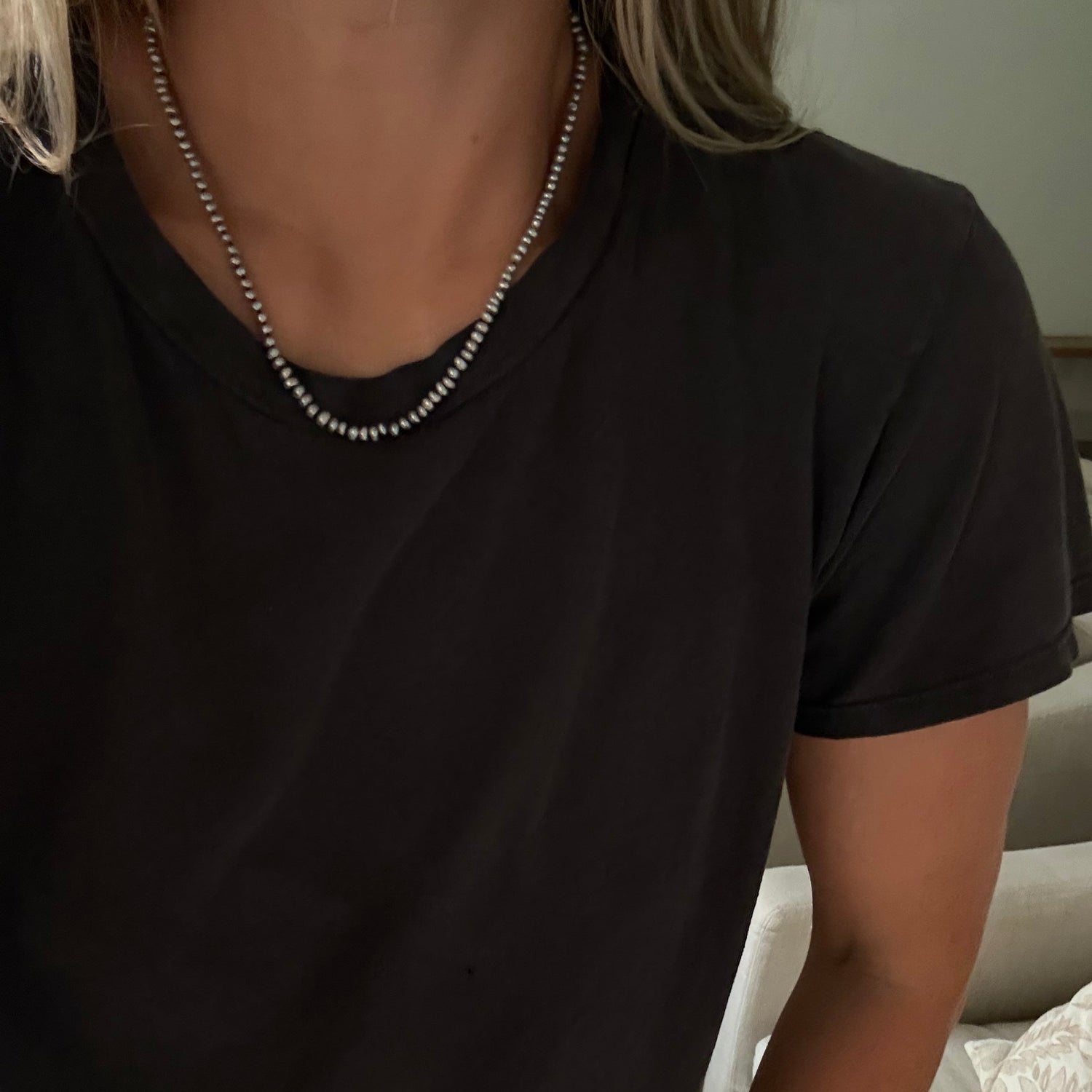 A close-up of a person wearing a gray freshwater pearl necklace  over a black t-shirt, with blonde hair visible at the top.