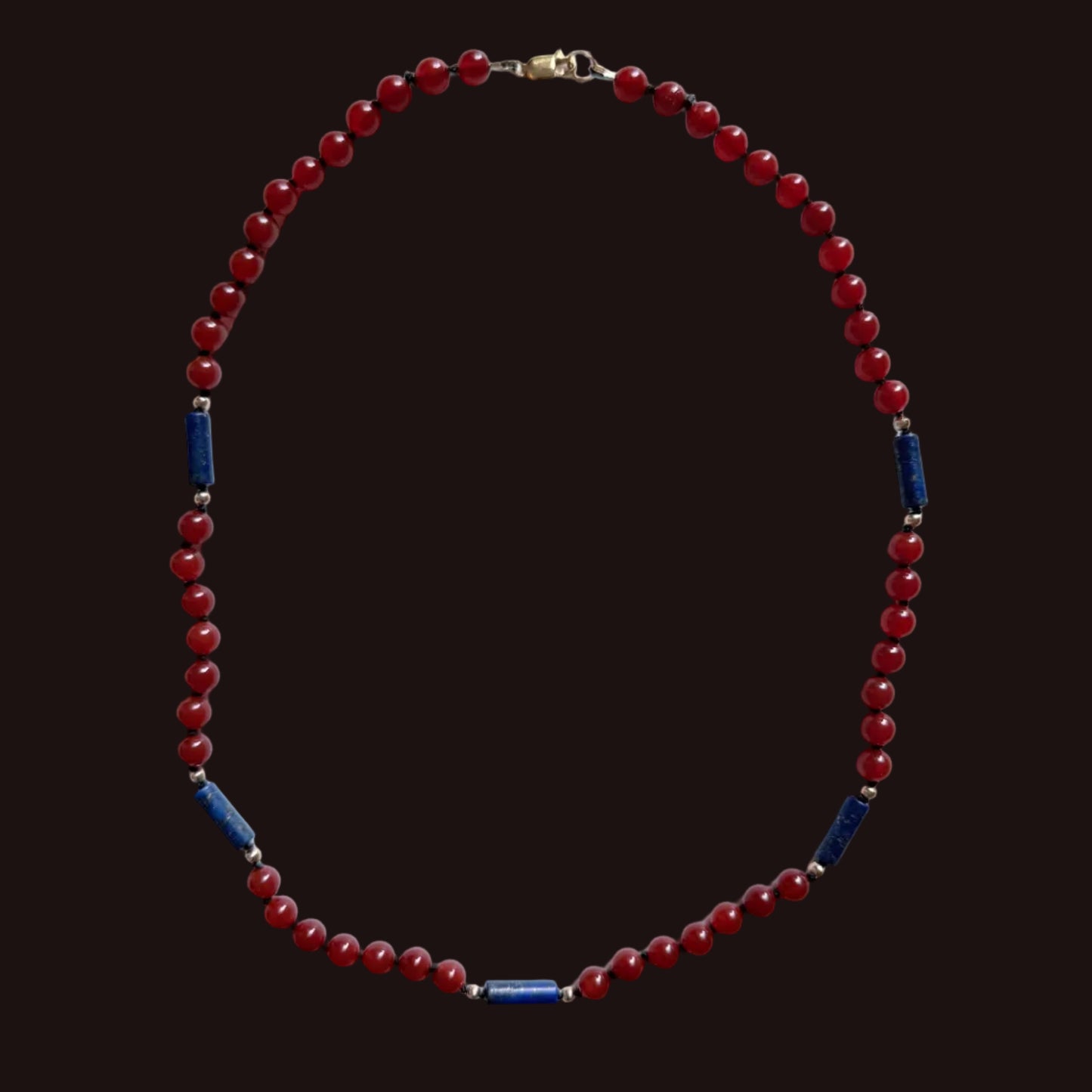 A beaded necklace featuring round red beads and alternating dark blue cylindrical spacer beads separated by small silver-toned metal rings, displayed on a light gray background.
