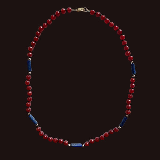 A beaded necklace featuring round red beads and alternating dark blue cylindrical spacer beads separated by small silver-toned metal rings, displayed on a light gray background.