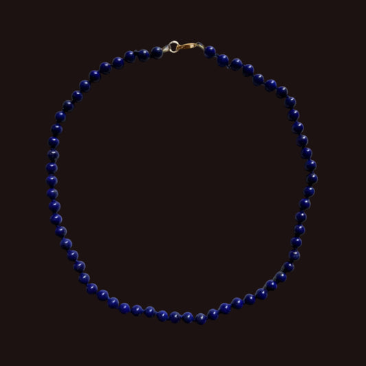 A beaded necklace made of small dark blue beads with a gold clasp, laid flat on a light gray background.