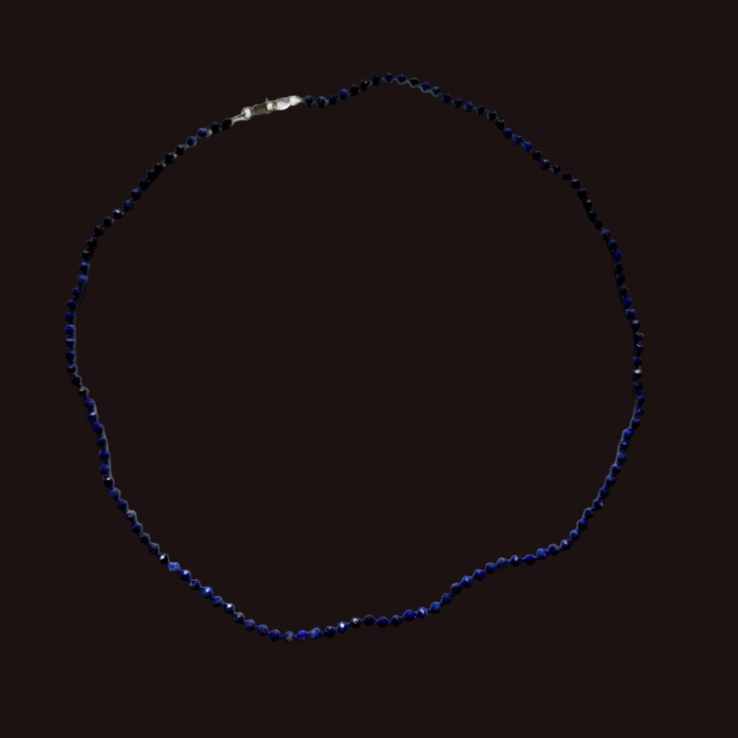 A beaded necklace made of small dark blue faceted beads with a silver clasp, laid out in a circle on a white sheet of paper.