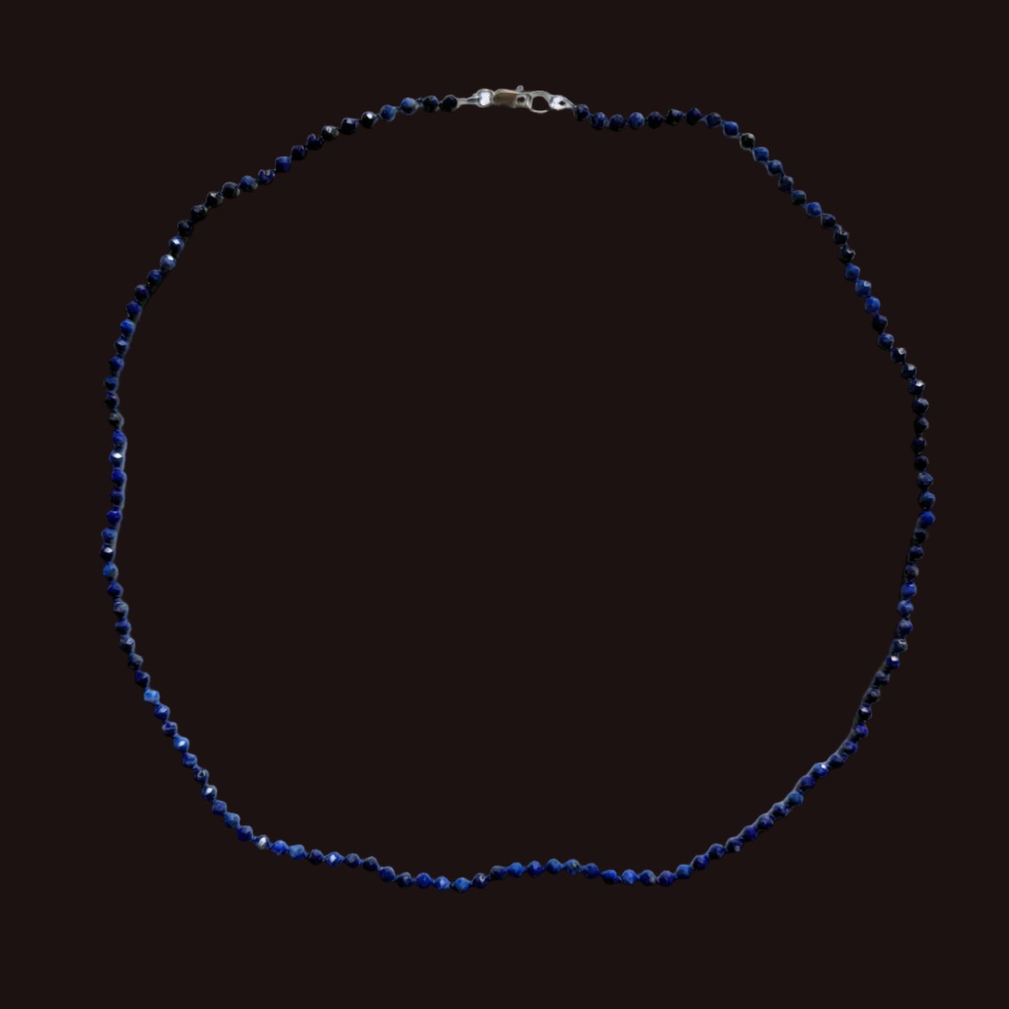 A beaded necklace made of small dark blue faceted beads with a silver clasp, arranged in a circle on a plain light gray background.
