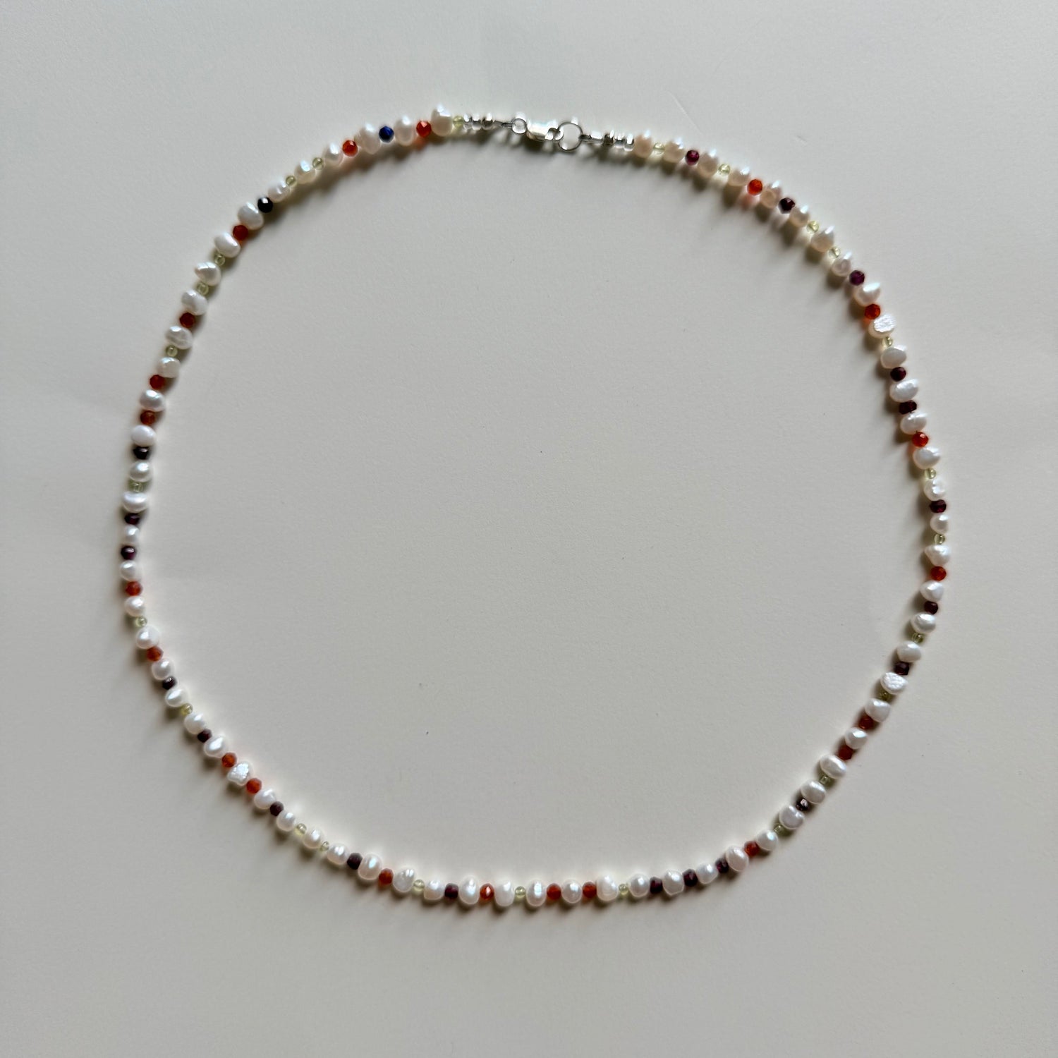 A beaded necklace made of small white, red, and dark brown beads with a silver clasp, laid flat on a light gray background.