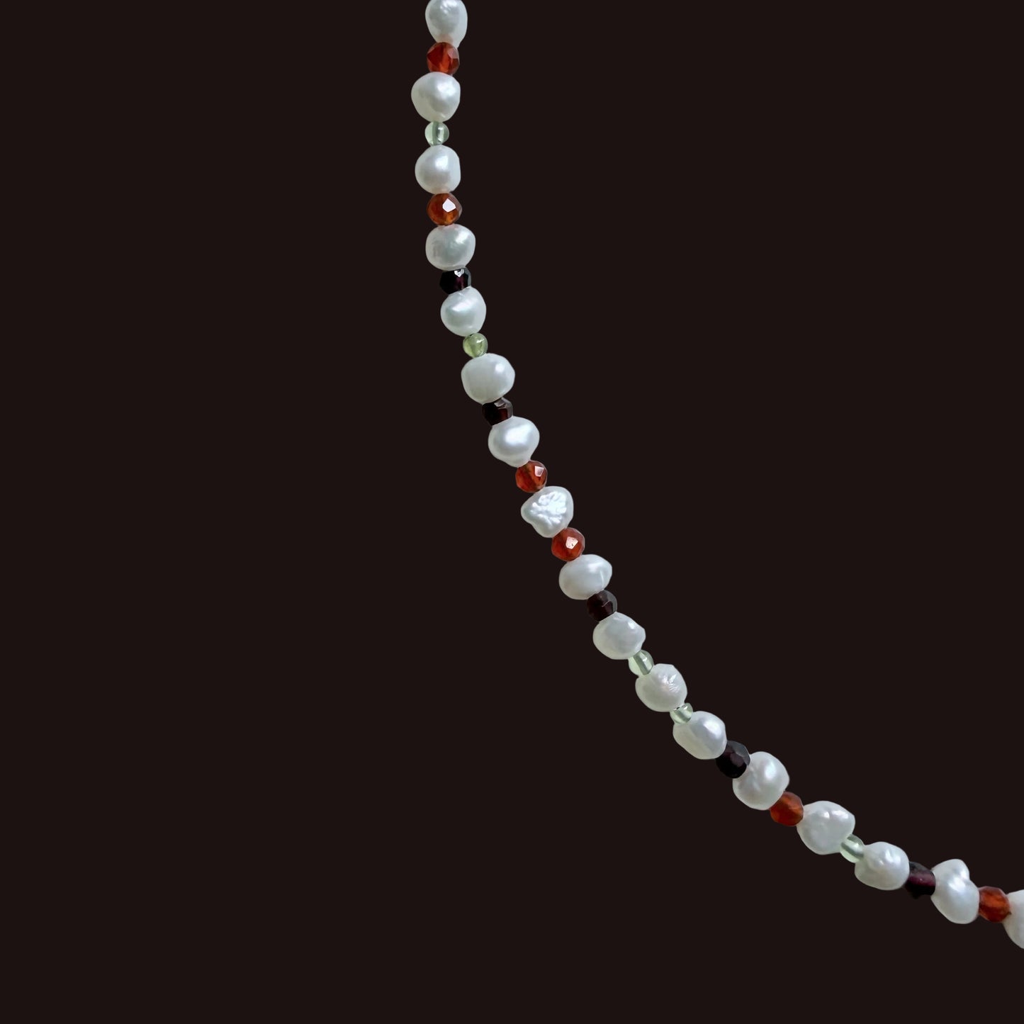 A beaded strand necklace featuring white baroque pearls and small faceted beads in red, dark brown, and light green, arranged diagonally on a light gray background.