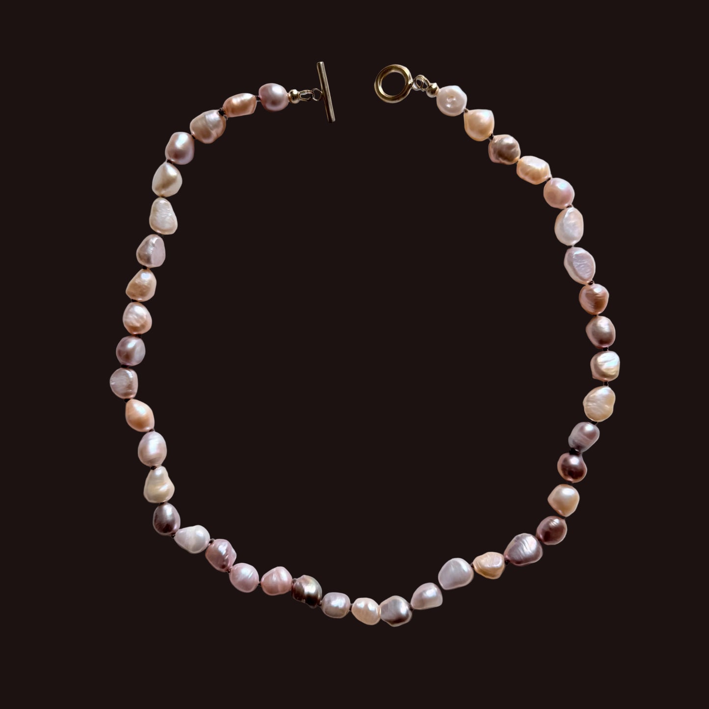 A beaded necklace made of irregular, baroque-style pearls in soft pink, peach, cream, and mauve tones, arranged in a circle on a light gray background. It features a gold-tone toggle clasp on one end and a lobster clasp on the other.