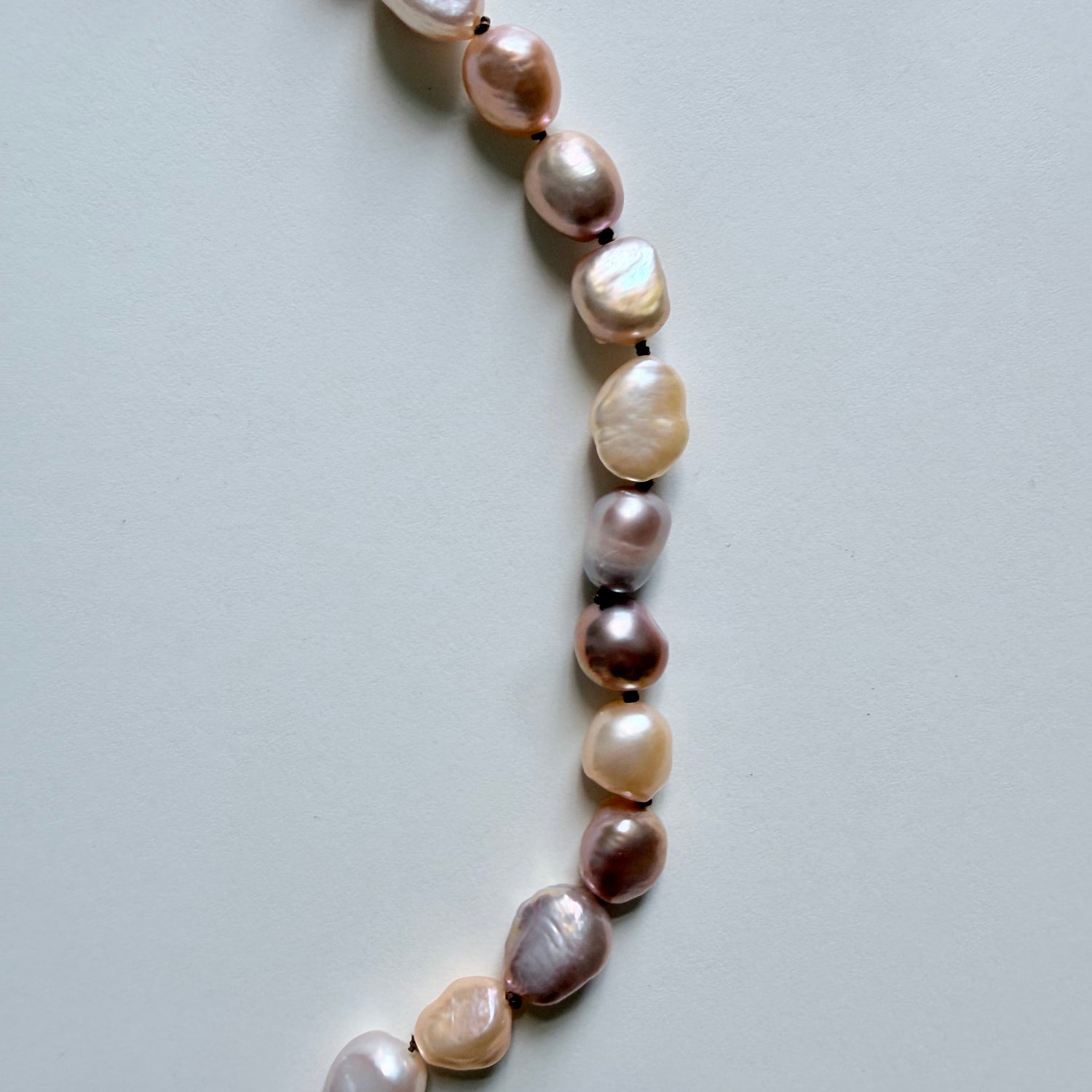 A close-up of a strand of irregular baroque pearls in soft champagne, beige, and dusty mauve-pink tones, laid out on a light gray background.