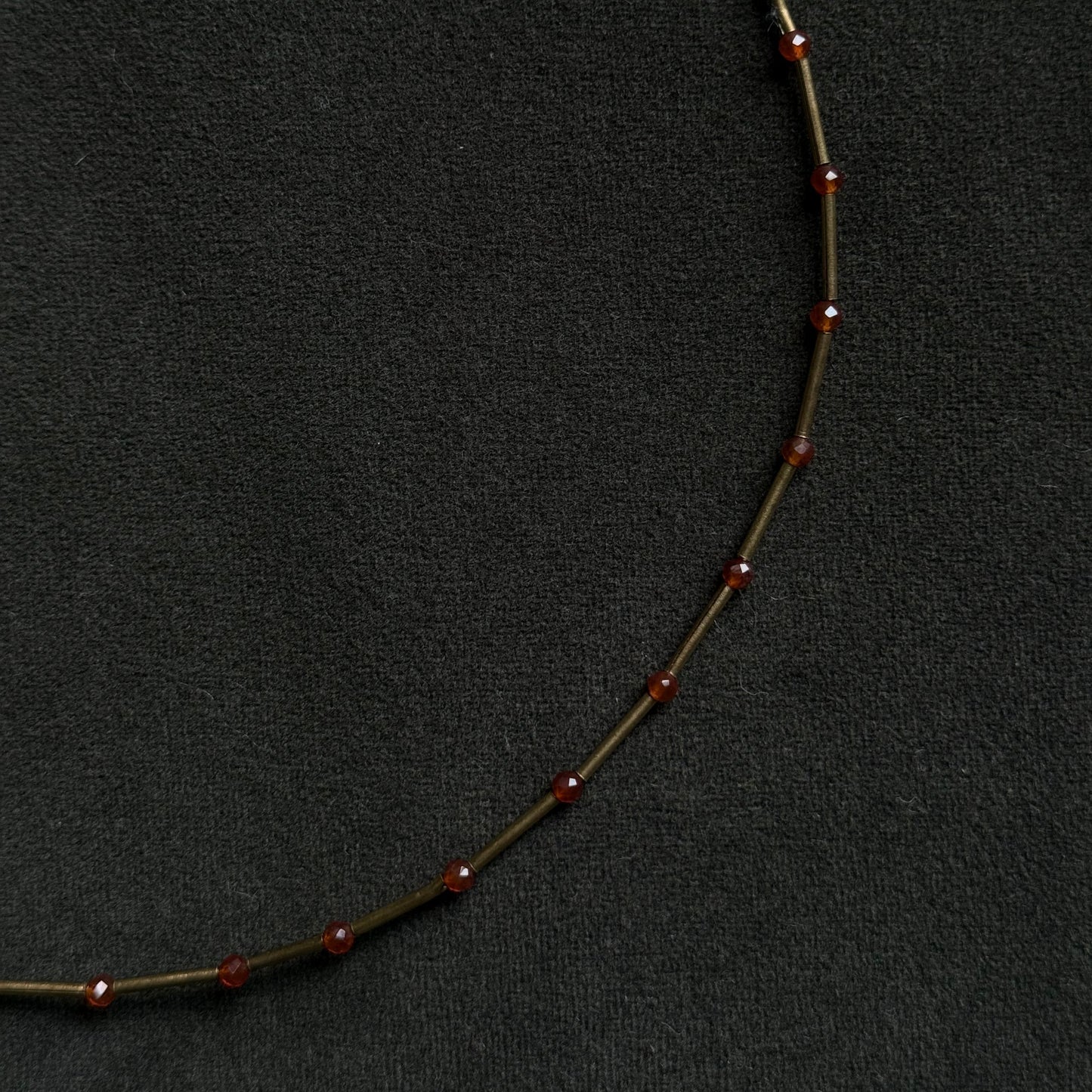 A close-up of a delicate necklace featuring small, round, dark red beads spaced along a thin, dark metallic chain, set against a textured black background.
