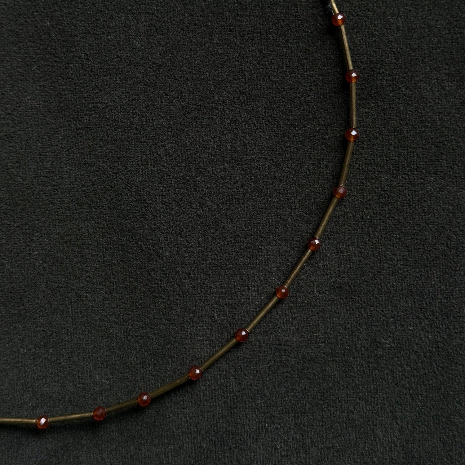 A close-up of a delicate necklace featuring small, round, dark red beads spaced along a thin, dark metallic chain, set against a textured black background.