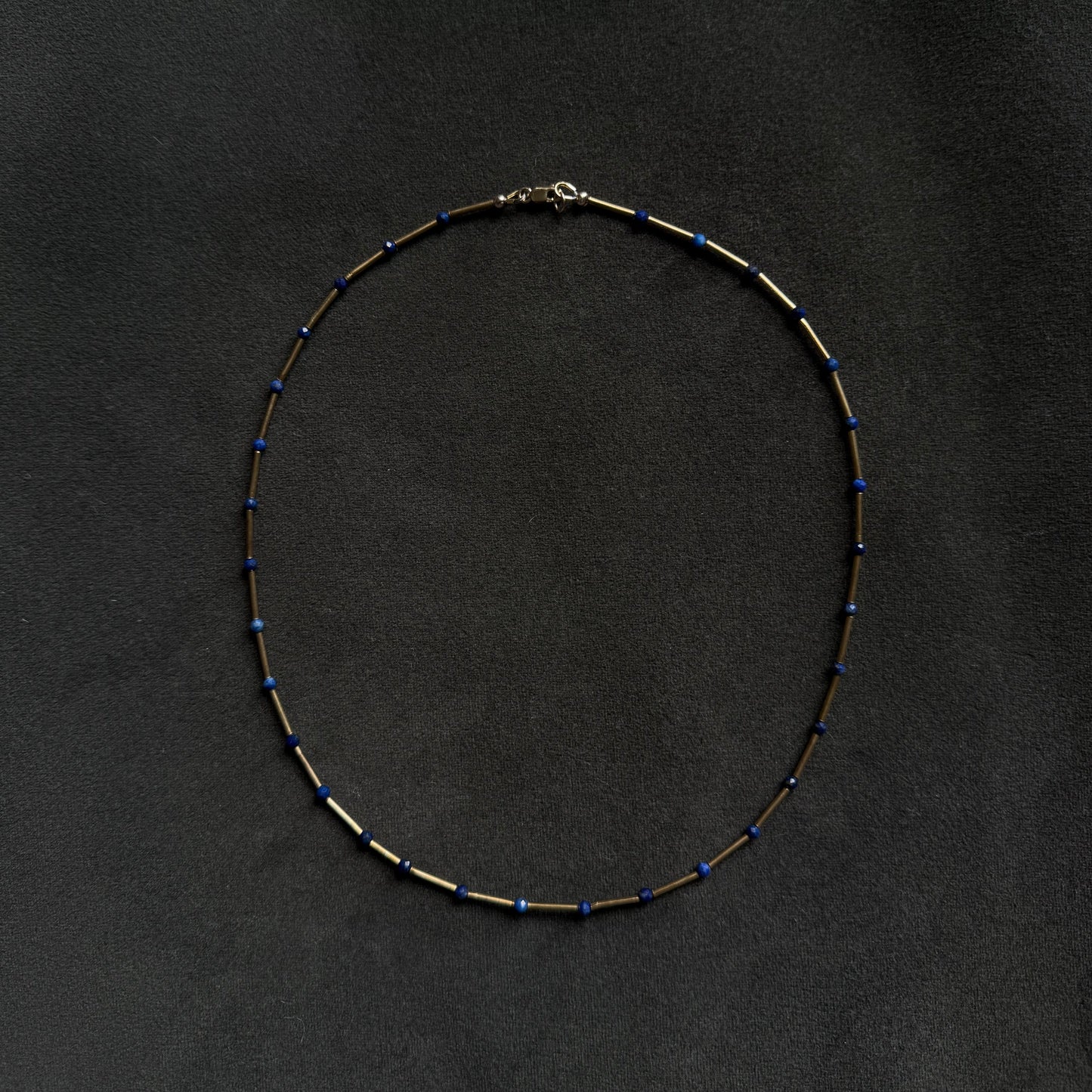 A delicate beaded necklace with small blue beads spaced along a dark metallic chain, laid flat on a dark textured background.