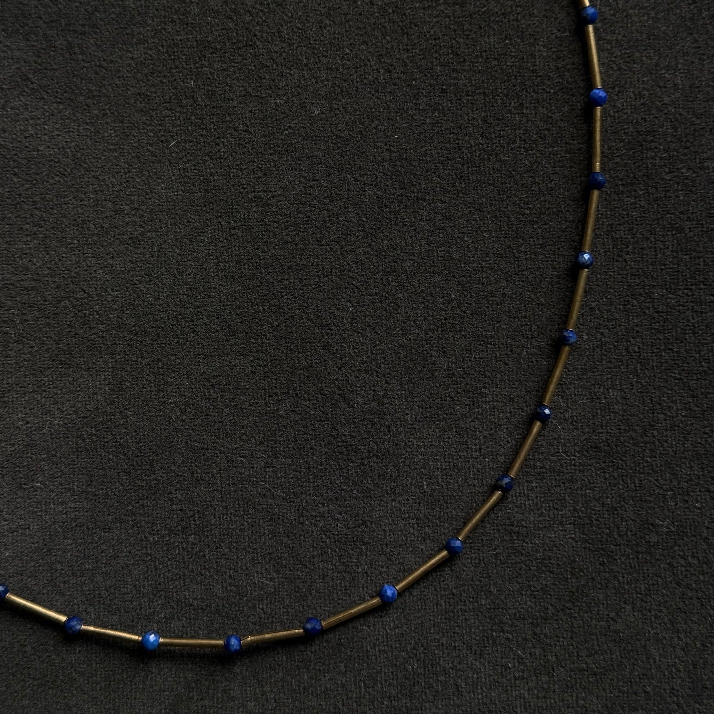 A delicate necklace made of small gold-toned tubes separated by tiny dark blue beads, laid in a curved shape on a dark textured background.