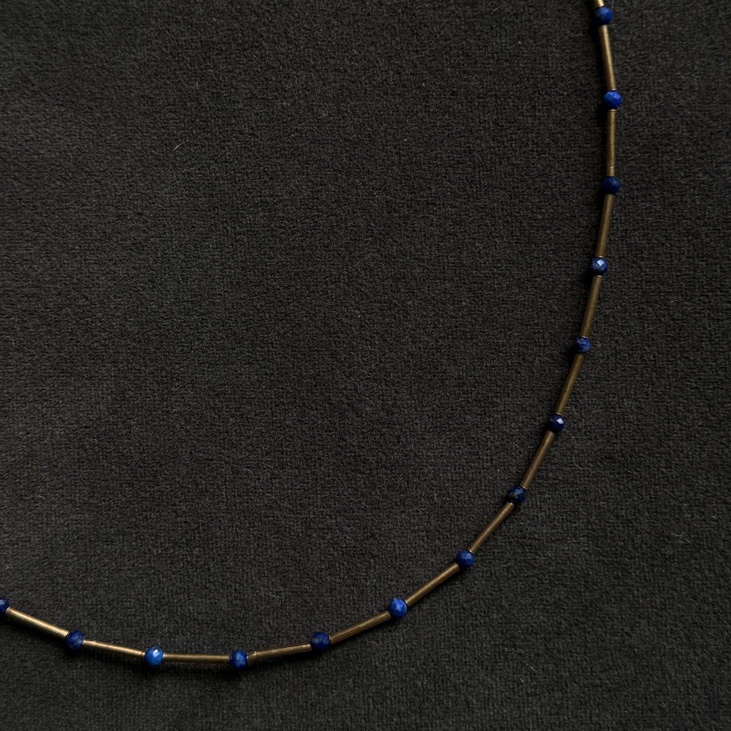 A delicate necklace made of small gold-toned tubes separated by tiny dark blue beads, laid in a curved shape on a dark textured background.