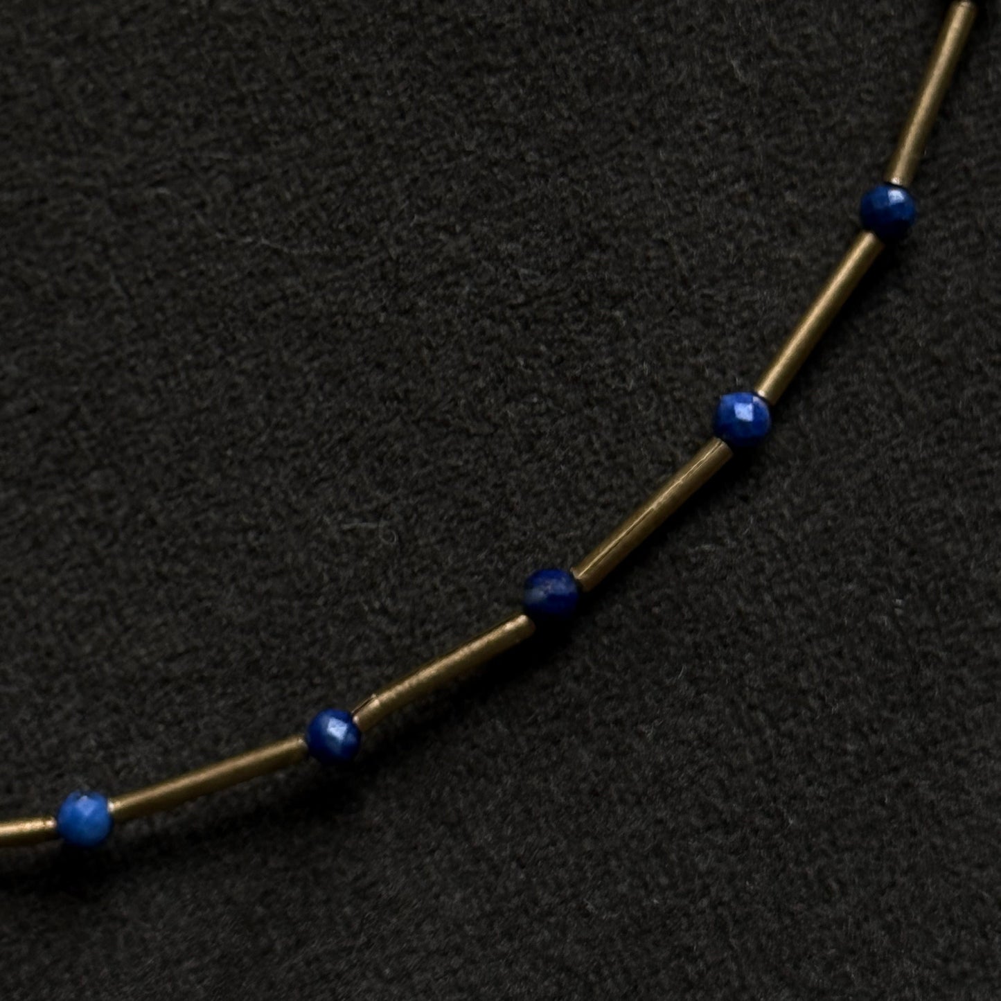A close-up of a necklace featuring small, faceted dark blue beads spaced along a thin, metallic gold-toned chain, set against a textured black background.
