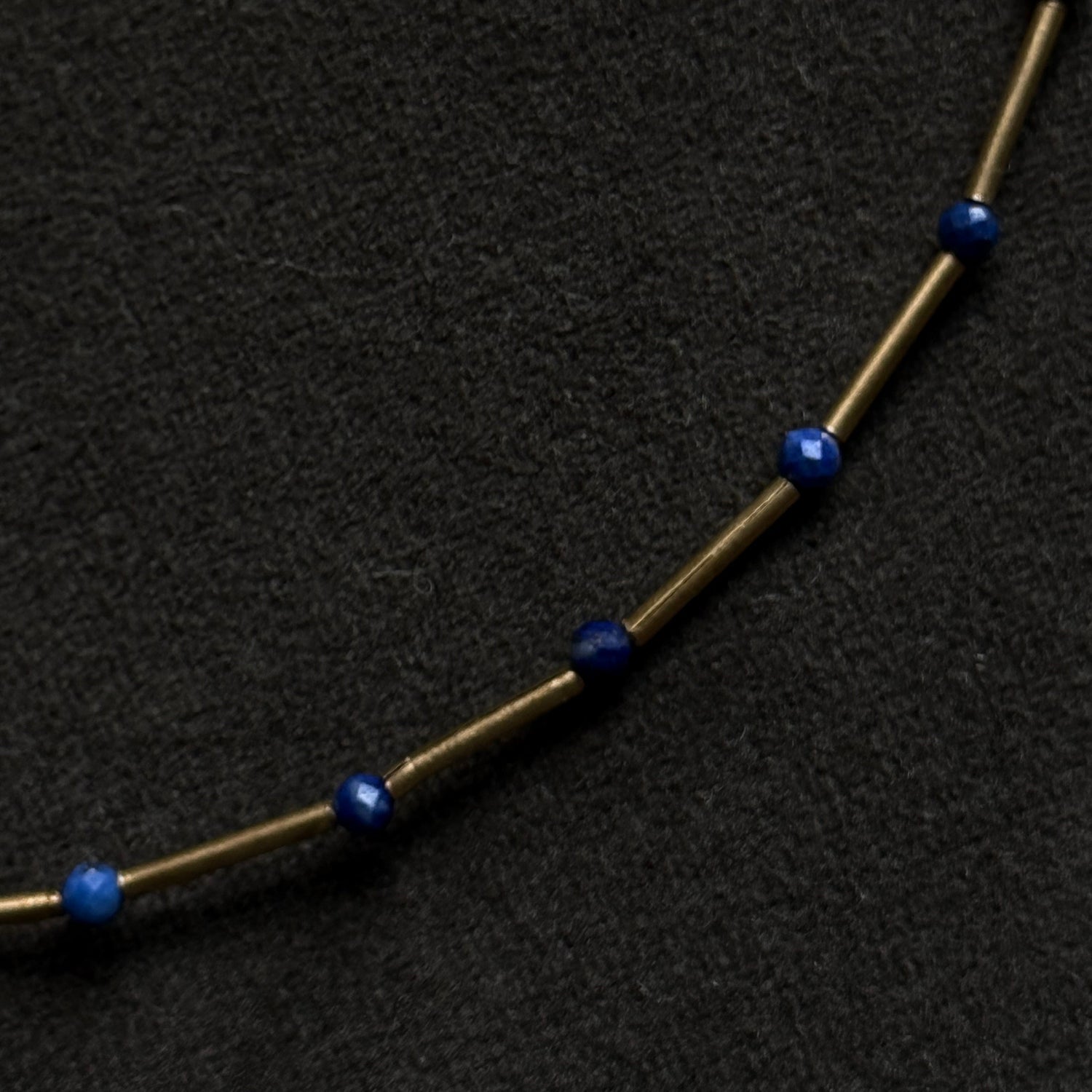 A close-up of a necklace featuring small, faceted dark blue beads spaced along a thin, metallic gold-toned chain, set against a textured black background.