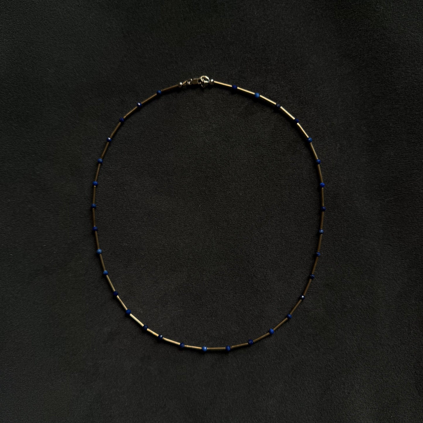 A delicate beaded necklace with alternating small gold and dark blue beads, arranged in a circle on a dark textured background.