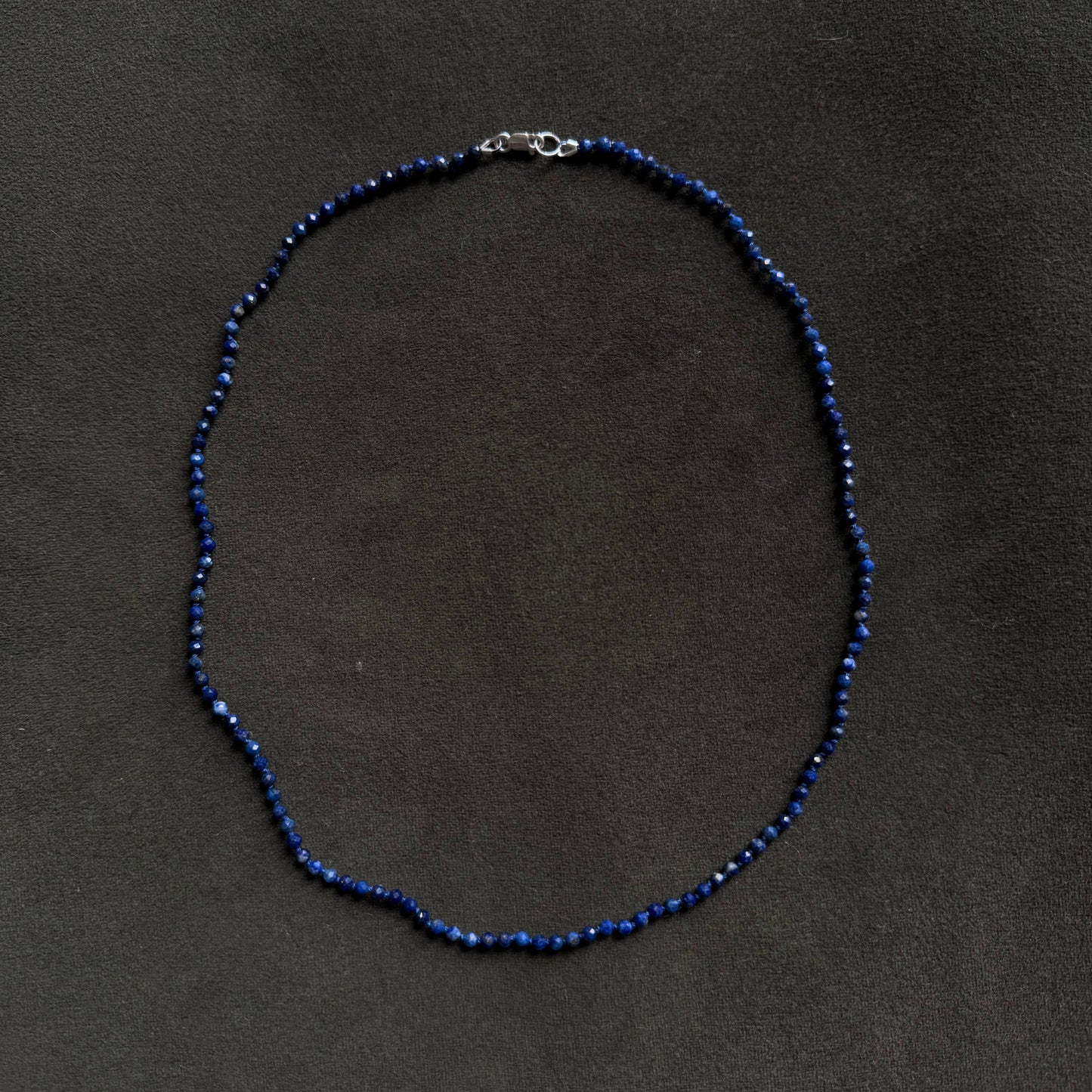 A beaded necklace made of small dark blue beads with a silver clasp, laid flat on a dark gray textured background.