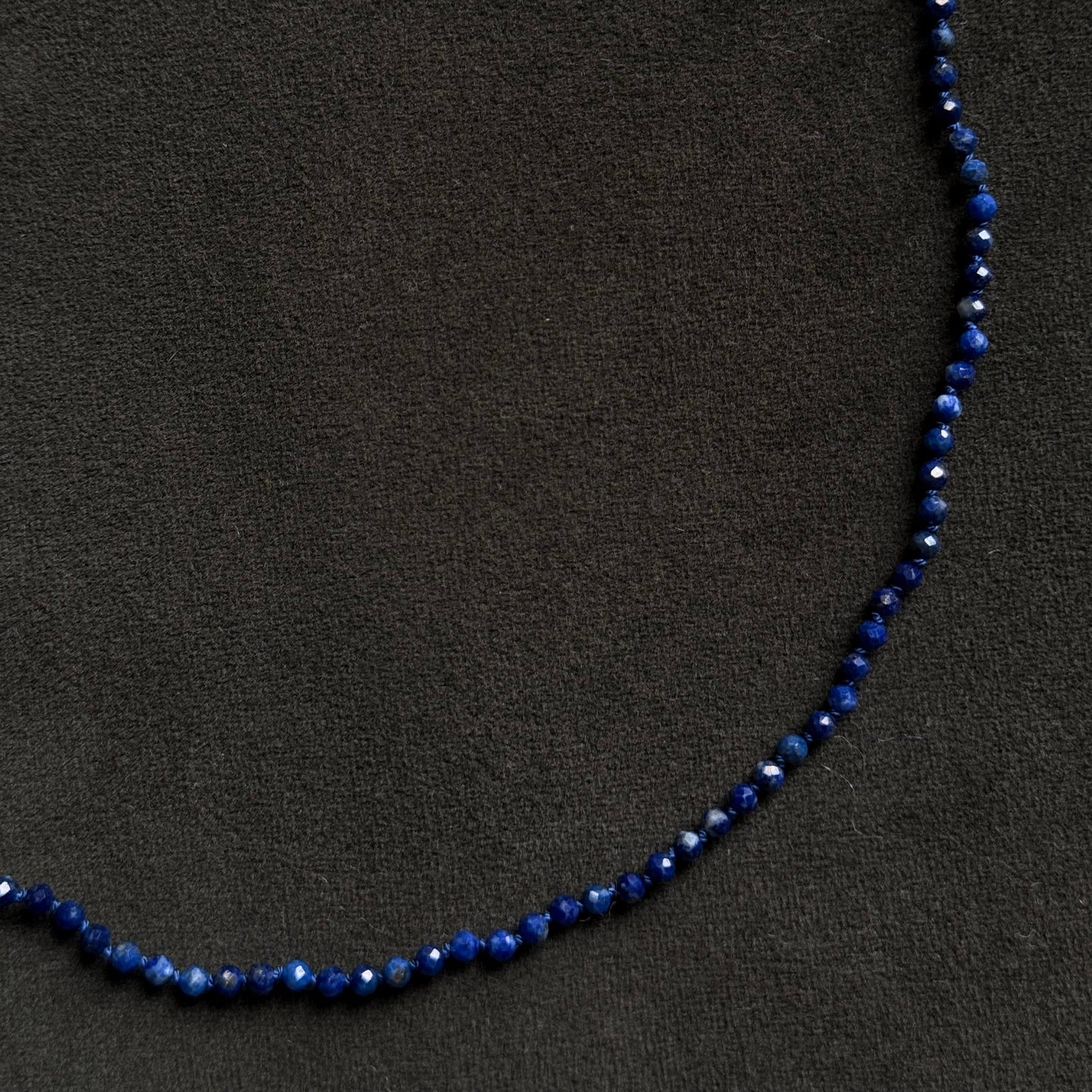 A beaded necklace made of small dark blue faceted beads, laid out in a curved shape on a dark textured background.