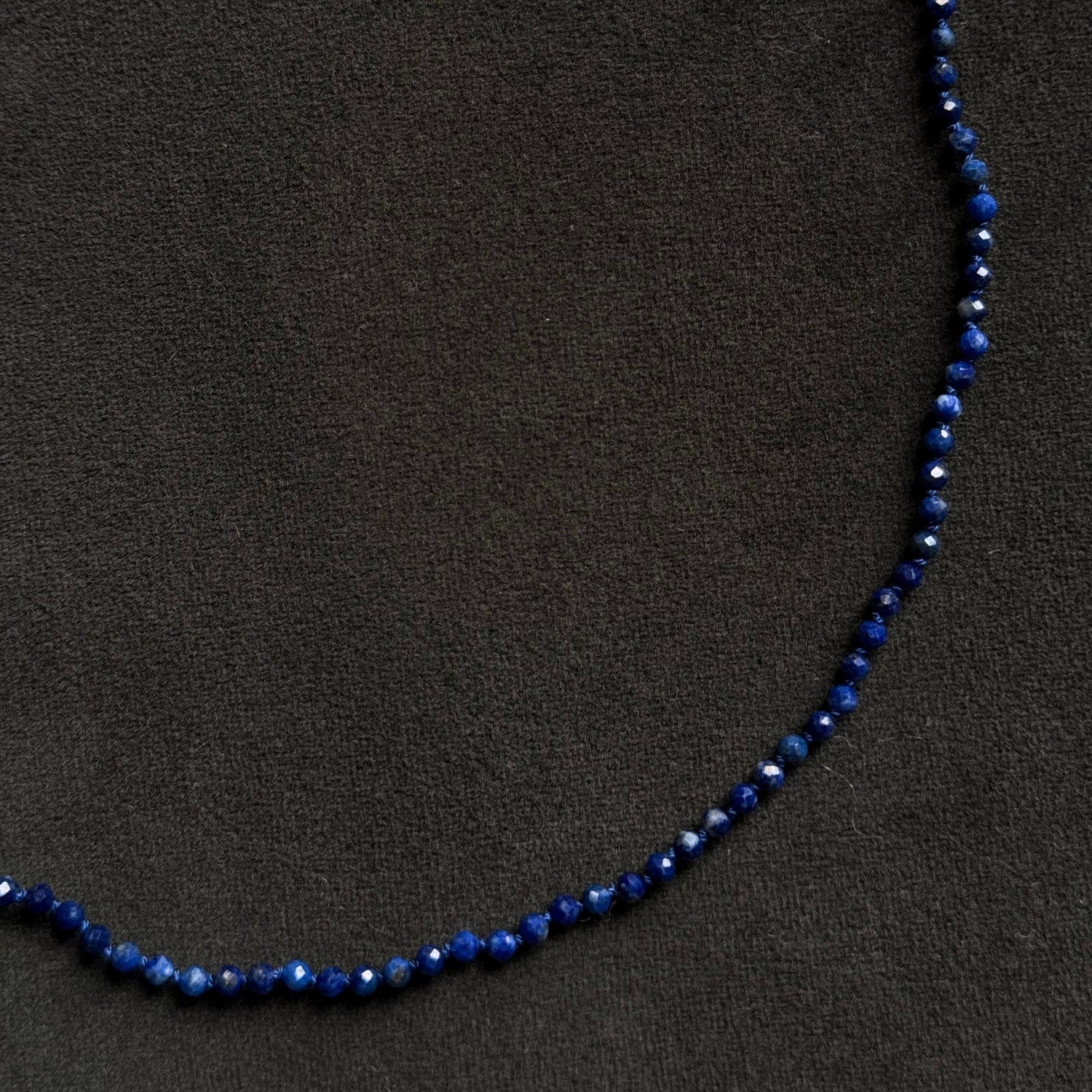 A beaded necklace made of small dark blue faceted beads, laid out in a curved shape on a dark textured background.