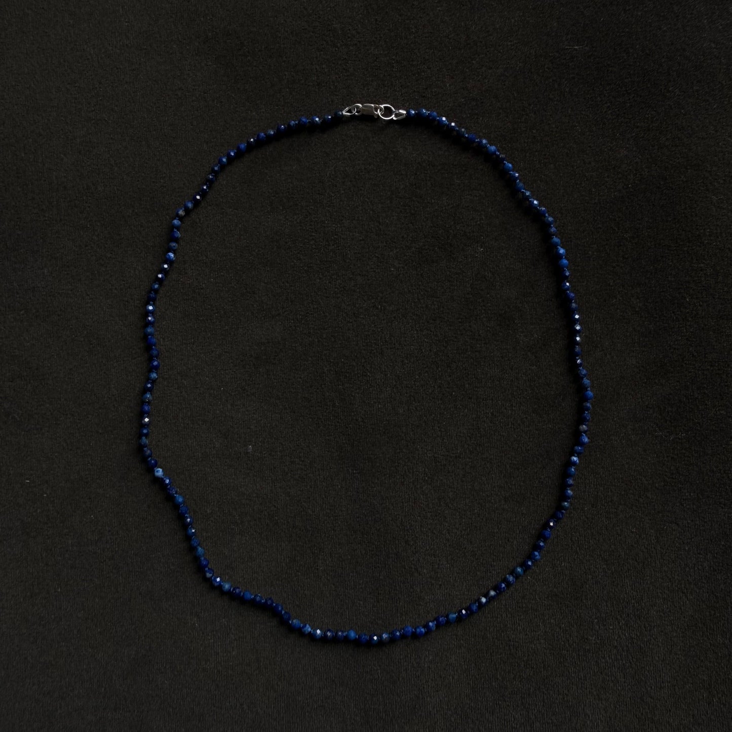 A beaded necklace made of small dark blue beads with a silver clasp, laid flat on a dark black background.