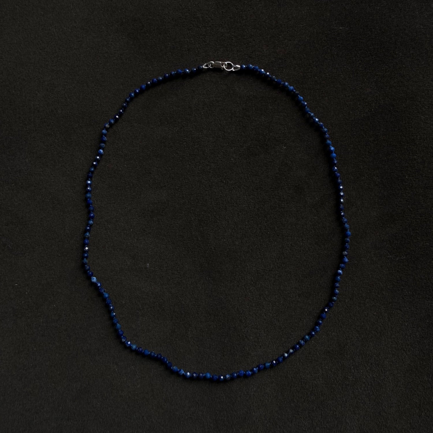 A beaded necklace made of small dark blue beads with a silver clasp, laid flat on a dark black background.
