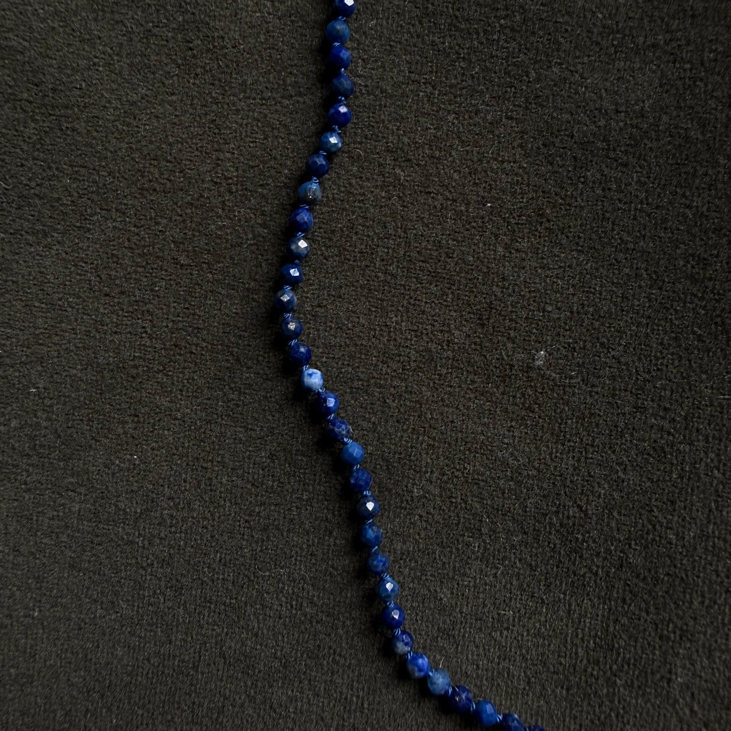 A strand of faceted dark blue beads, likely lapis lazuli, laid out diagonally on a textured black fabric background.