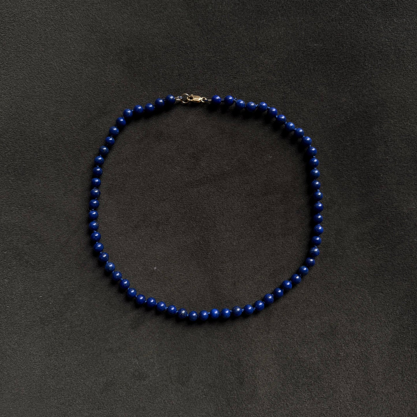 A beaded necklace made of small, glossy dark blue beads with a small gold clasp, displayed on a dark textured background.
