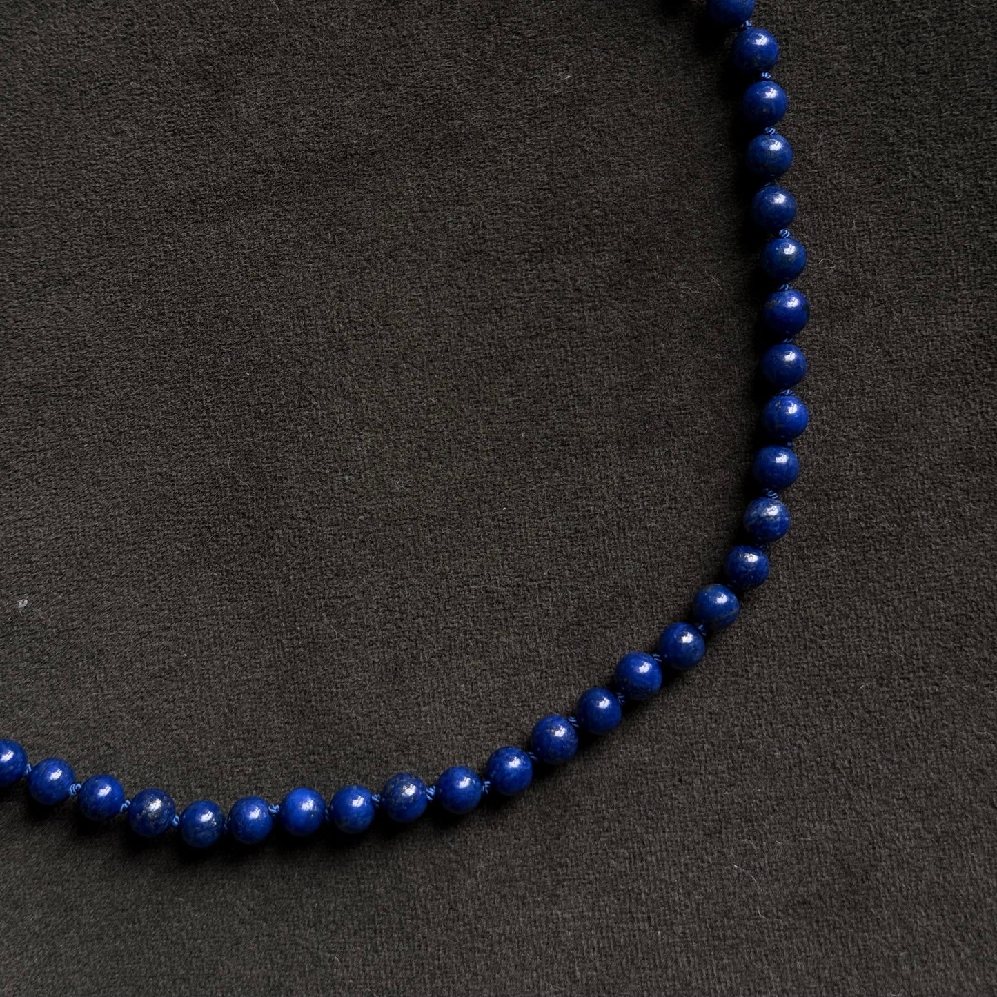 A beaded necklace made of polished dark blue lapis lazuli stones, arranged in a circle on a dark textured background. A gold-toned lobster clasp is visible at the top left.