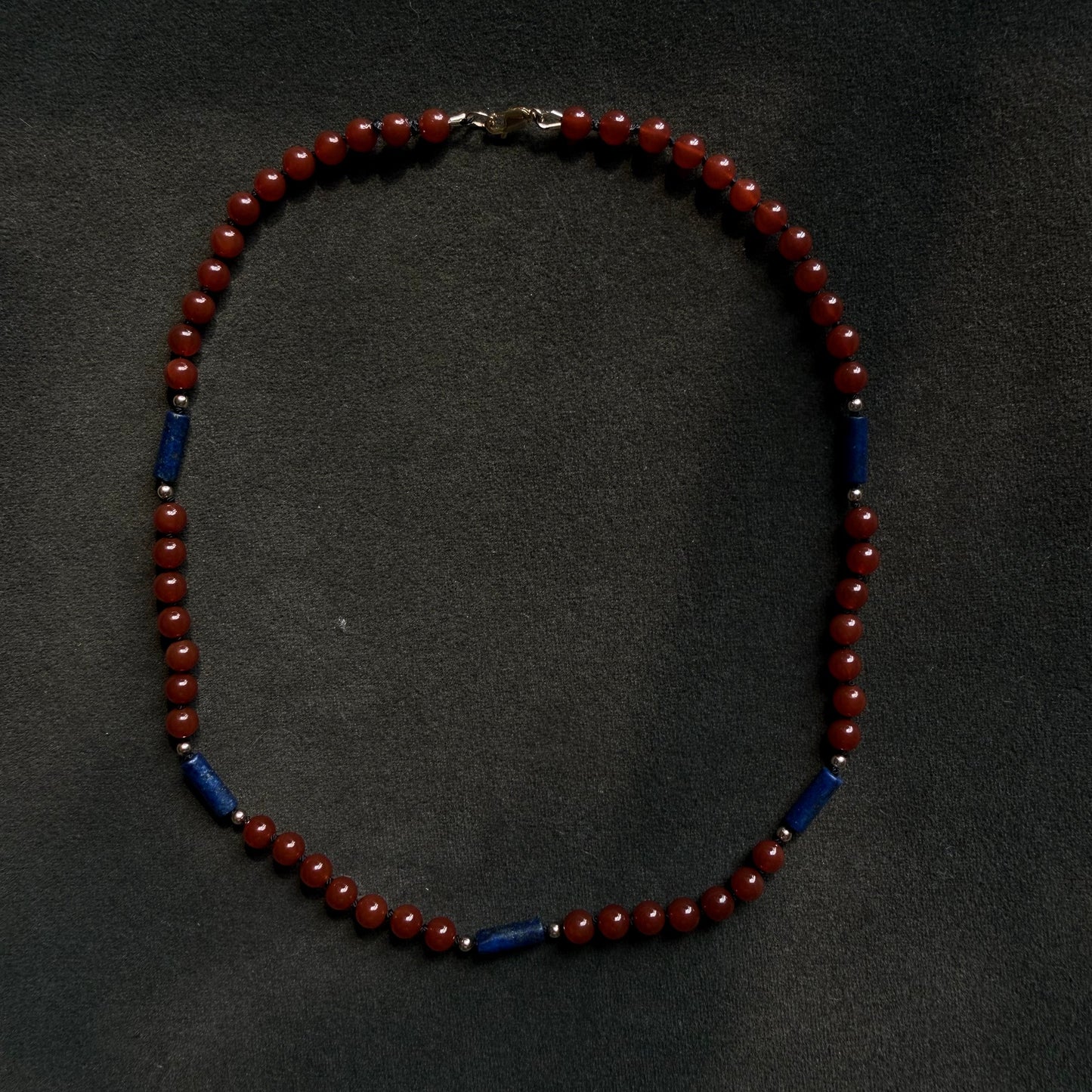 A beaded necklace with alternating deep red round beads and dark blue cylindrical beads, displayed on a dark textured background.