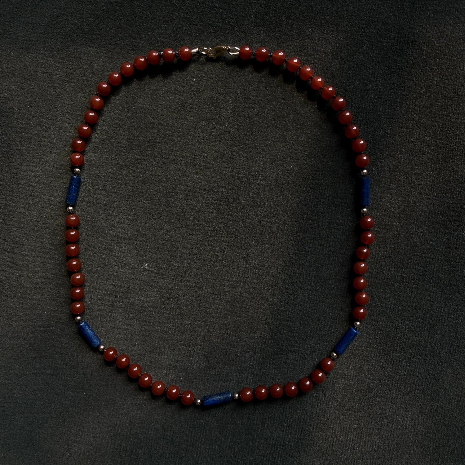 A beaded necklace with alternating deep red round beads and dark blue cylindrical beads, displayed on a dark textured background.