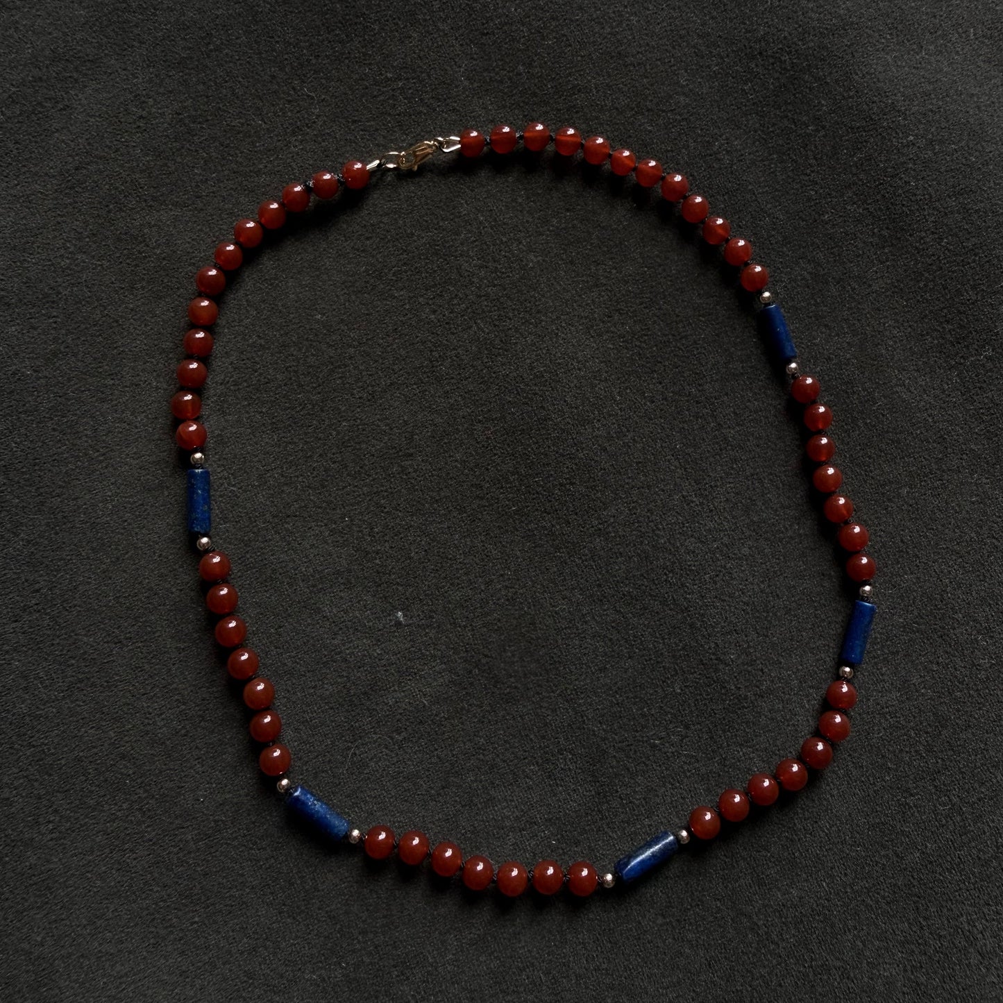 A beaded necklace featuring round red beads separated by small blue cylindrical spacers, displayed on a dark textured background.