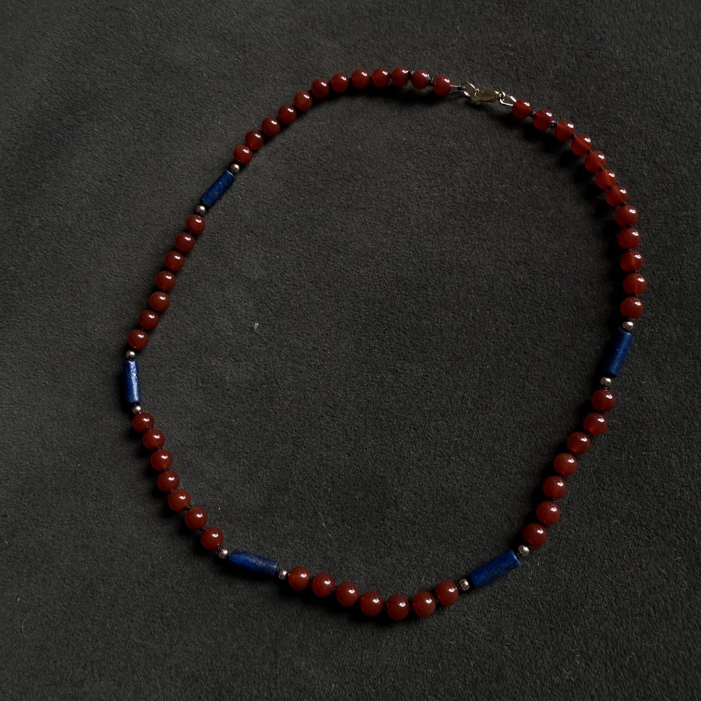 A beaded necklace with small round red beads and alternating dark blue cylindrical spacer beads, displayed on a dark textured background.