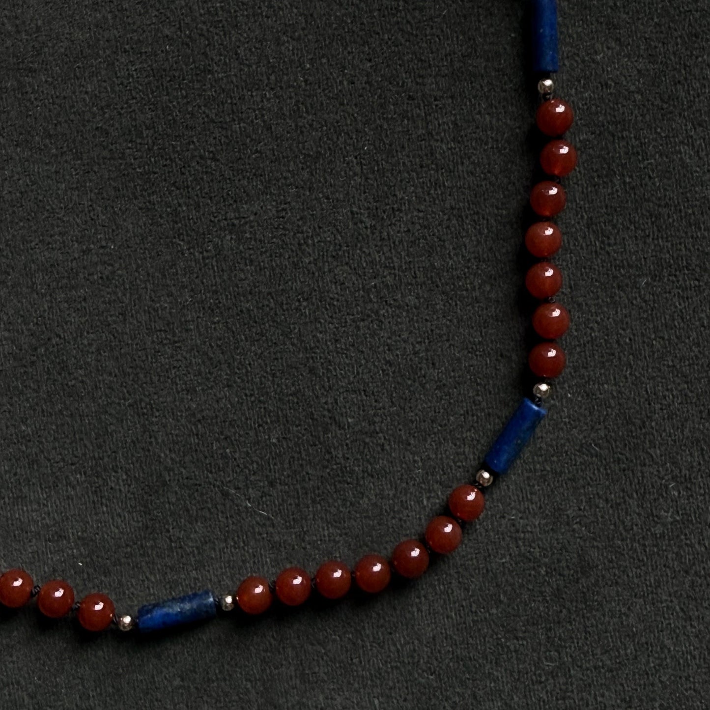 A beaded necklace featuring round red beads and two blue cylindrical spacer beads, displayed on a dark textured background.