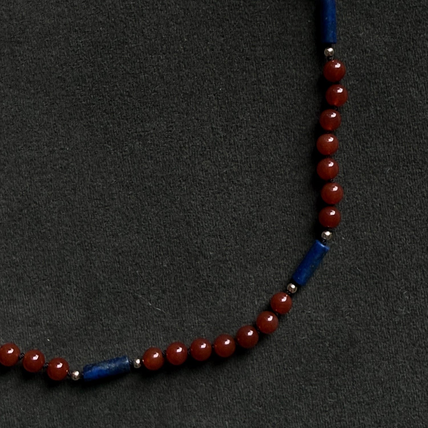A beaded necklace featuring round red beads and two blue cylindrical spacer beads, displayed on a dark textured background.