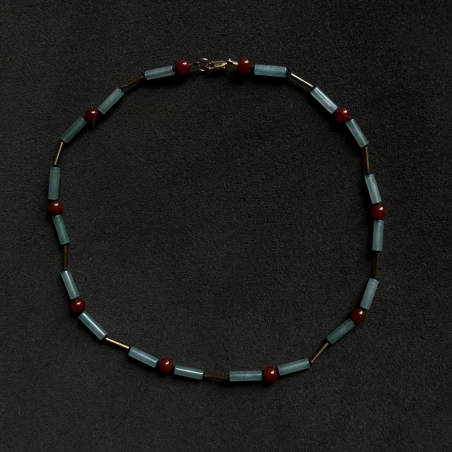 A beaded necklace featuring light blue-green rectangular beads separated by small dark brown cylindrical beads and accented with round dark red beads, displayed on a dark textured background.